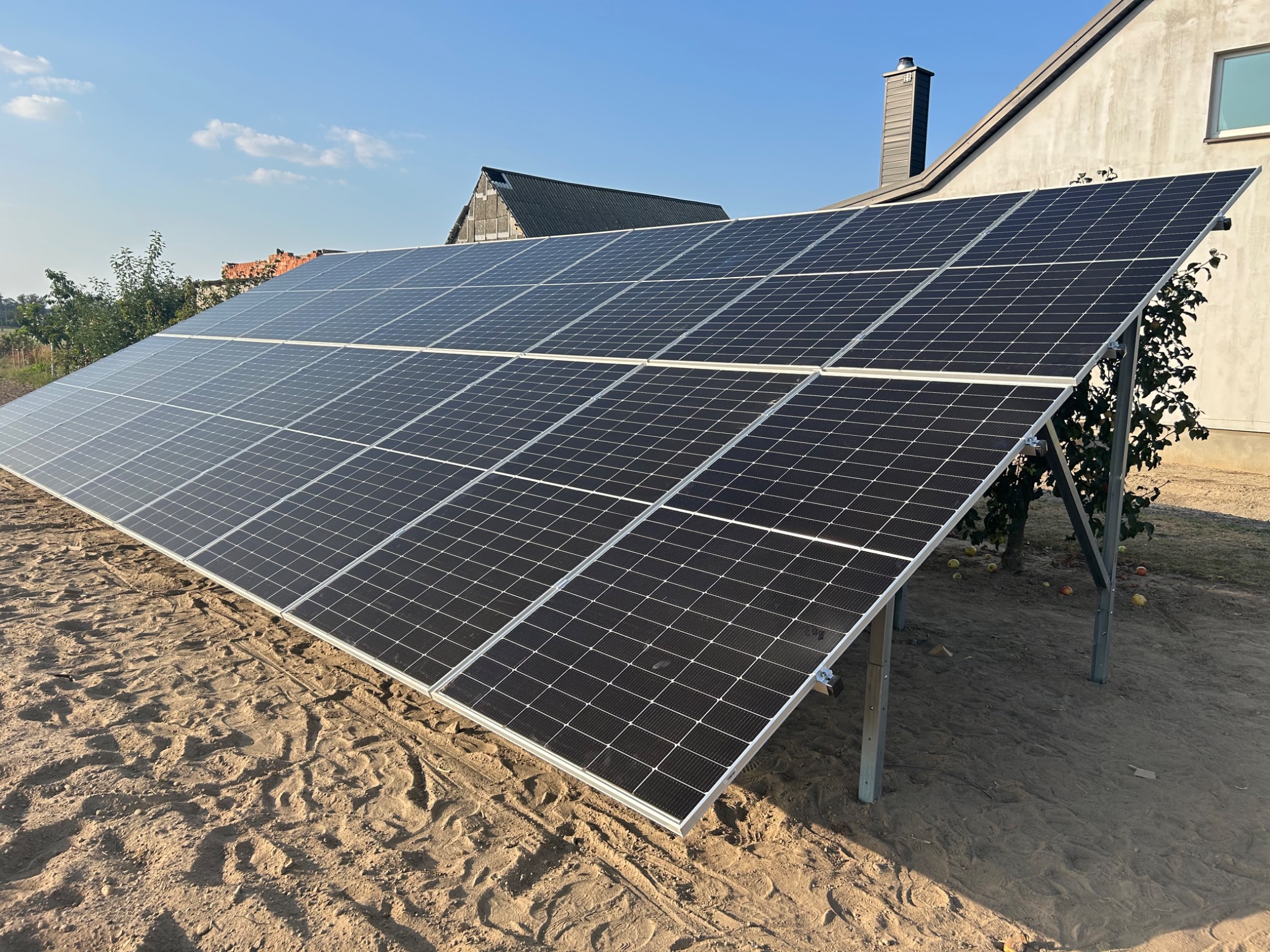 Instalacja fotowoltaiczna na gruncie z widocznymi panelami, metalową konstrukcją wsporczą i budynkiem w tle, w słoneczny dzień. Widoczne ślady opon w piasku.