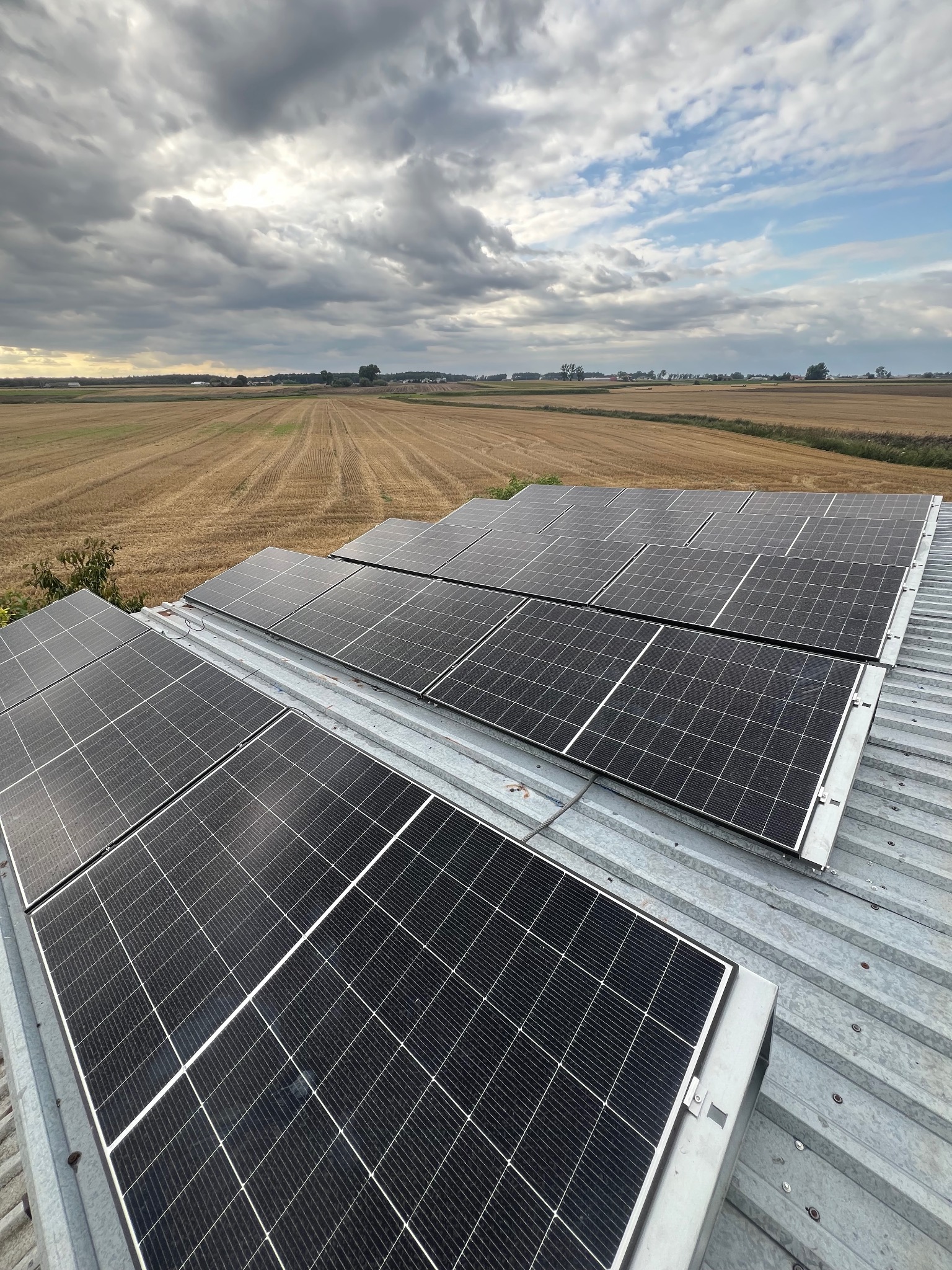Panele słoneczne na dachu z blachy falistej w Aleksandrowie Kujawskim, widok na pole i zachmurzone niebo. Montaż z bliska, widoczne detale mocowania.