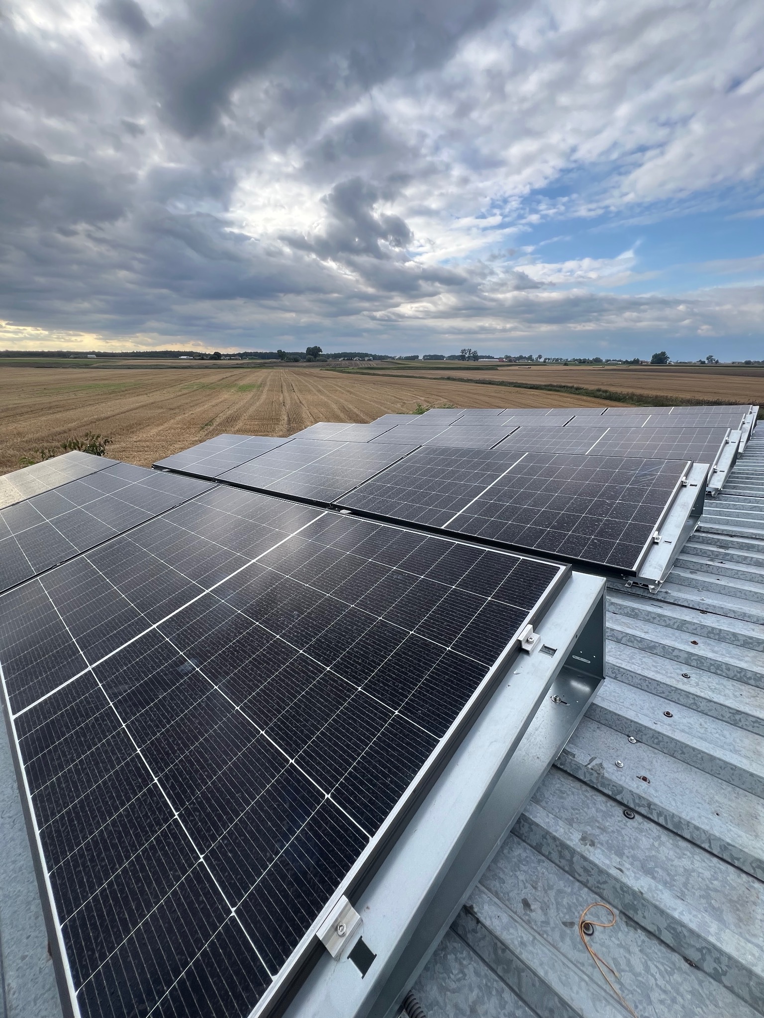 Rząd paneli fotowoltaicznych na metalowym dachu, z polami uprawnymi i pochmurnym niebem w tle. Widoczne elementy montażowe systemu.