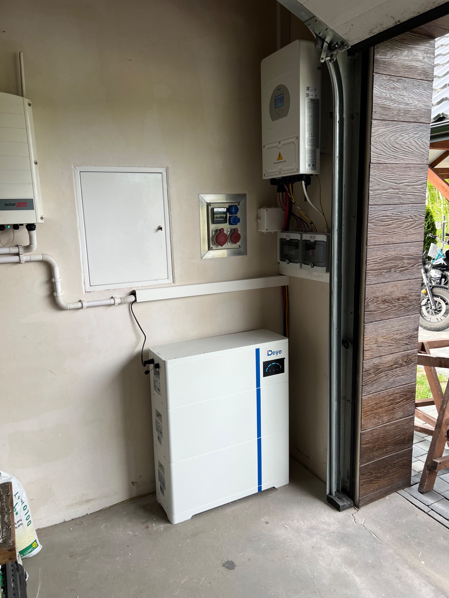 Wnętrze garażu z zamontowanym magazynem energii Deye, inwerterem SolarEdge i rozdzielnicą elektryczną. Widoczne przewody i instalacje. Częściowo widoczny motocykl na zewnątrz.