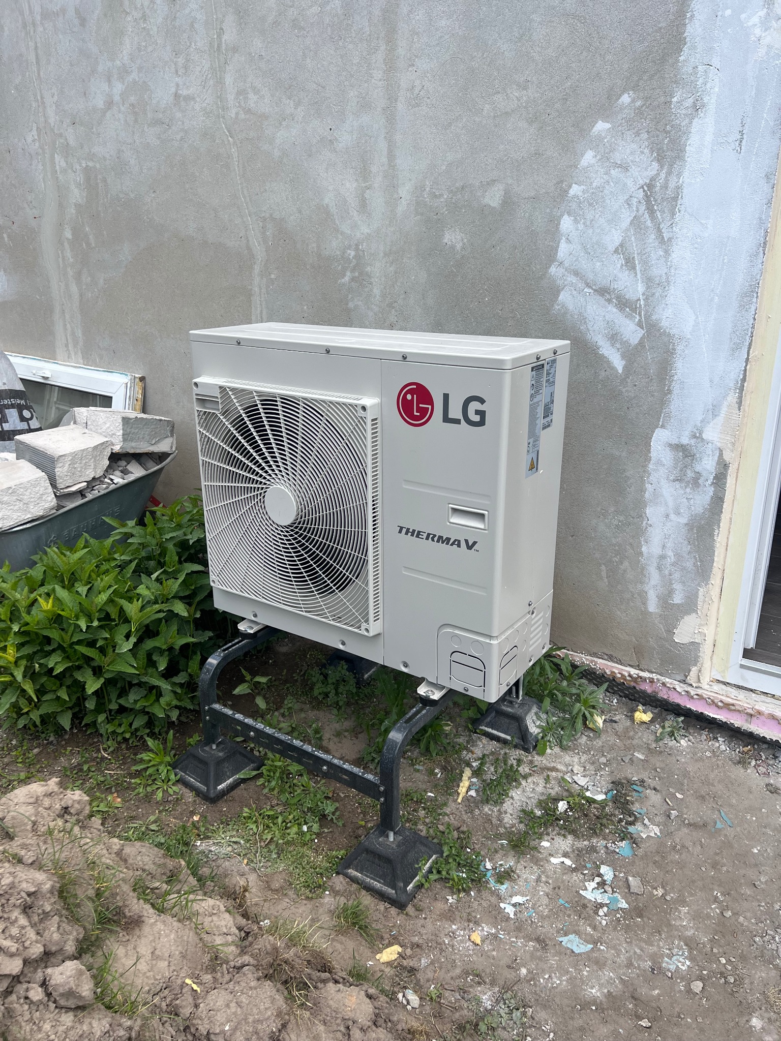 Biała pompa ciepła LG Therma V, zamontowana na czarnej konstrukcji, stoi przed surową ścianą budynku. Obok widoczne rośliny, taczka z kamieniami i okno.