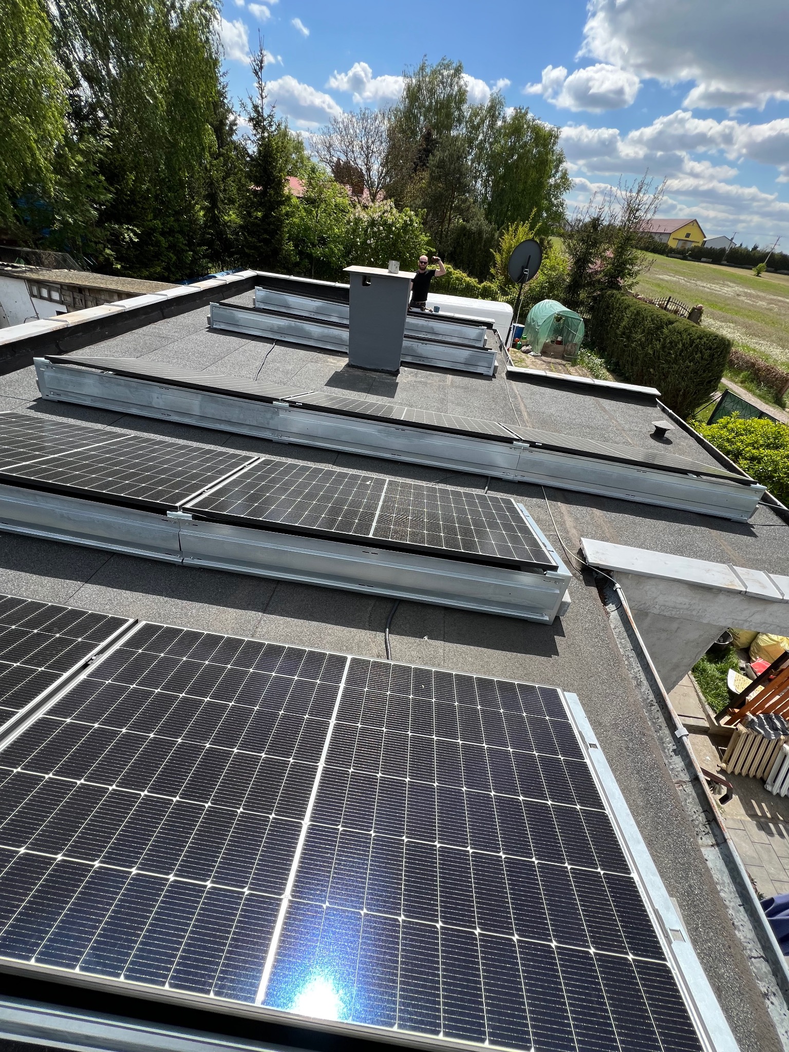 Instalacja fotowoltaiczna na płaskim dachu budynku jednorodzinnego, widok z góry, panele w metalowych ramach, w tle zieleń drzew i błękitne niebo.