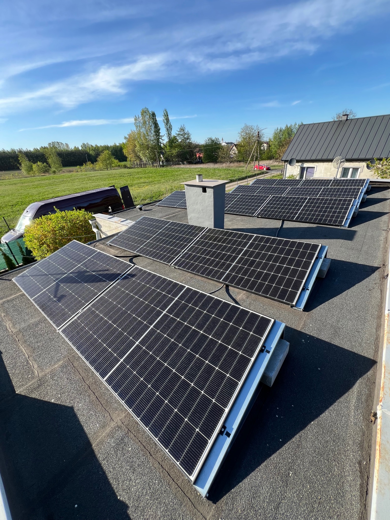 Panele fotowoltaiczne na dachu budynku z kominem, widoczne pole i zabudowania w tle, słoneczny dzień.