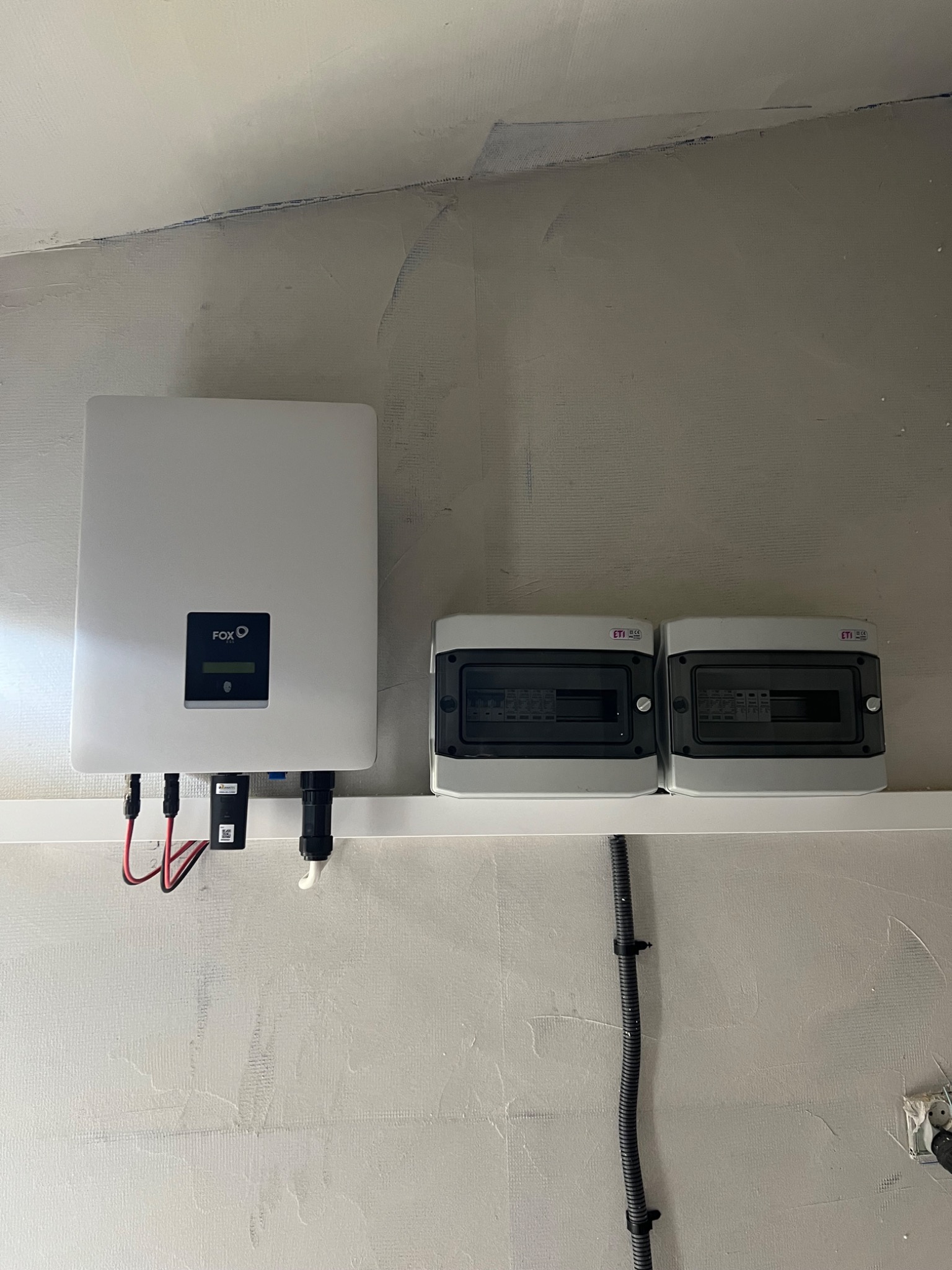 Biały falownik FoxESS z podłączonymi przewodami i dwie rozdzielnice ETI zamontowane na ścianie o surowym wykończeniu, widok z dołu na instalację.