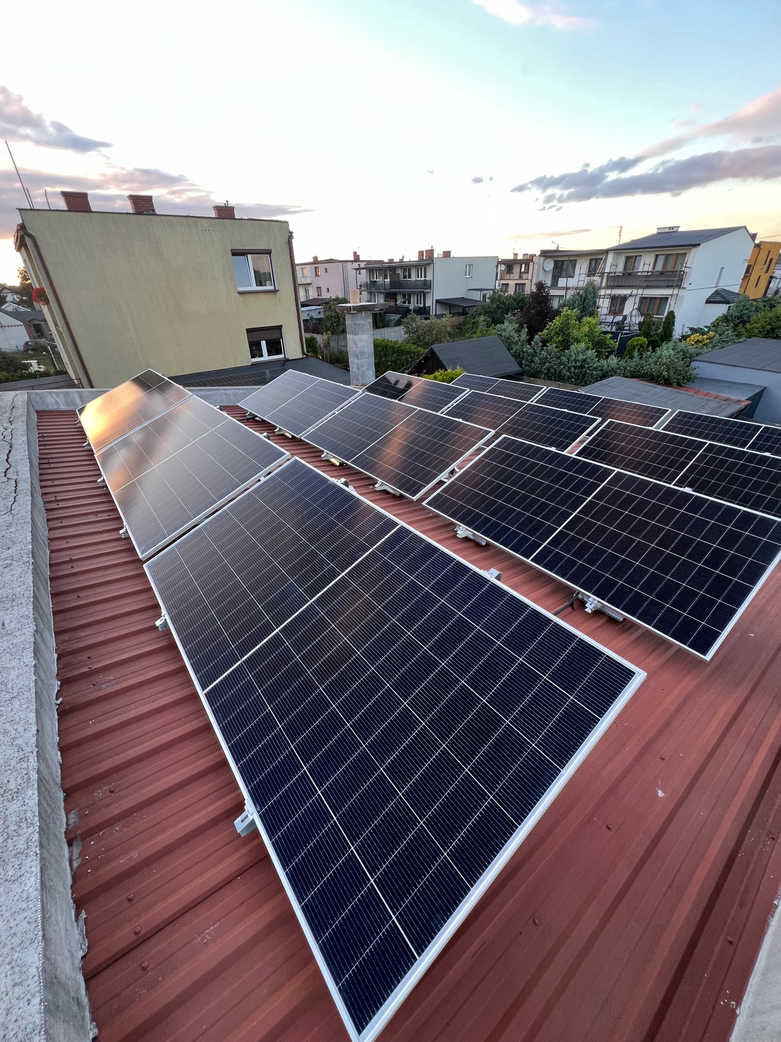 Panele fotowoltaiczne na czerwonym dachu, widok z góry, w tle zabudowa mieszkalna i zachodzące słońce odbijające się w panelach.