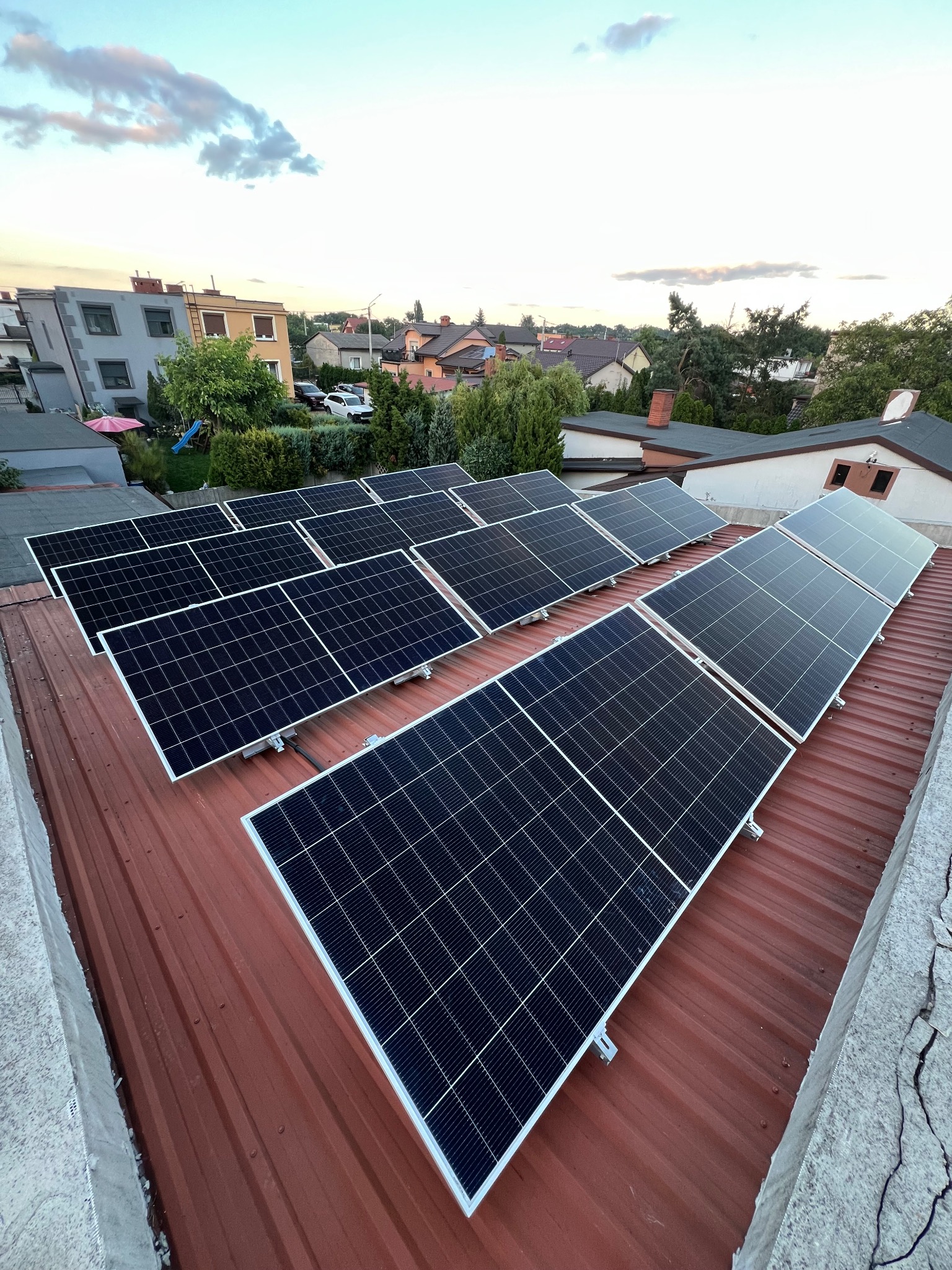 Rząd paneli fotowoltaicznych na czerwonym dachu z blachy, widok z góry, w tle zabudowa jednorodzinna i błękitne niebo o zmierzchu.