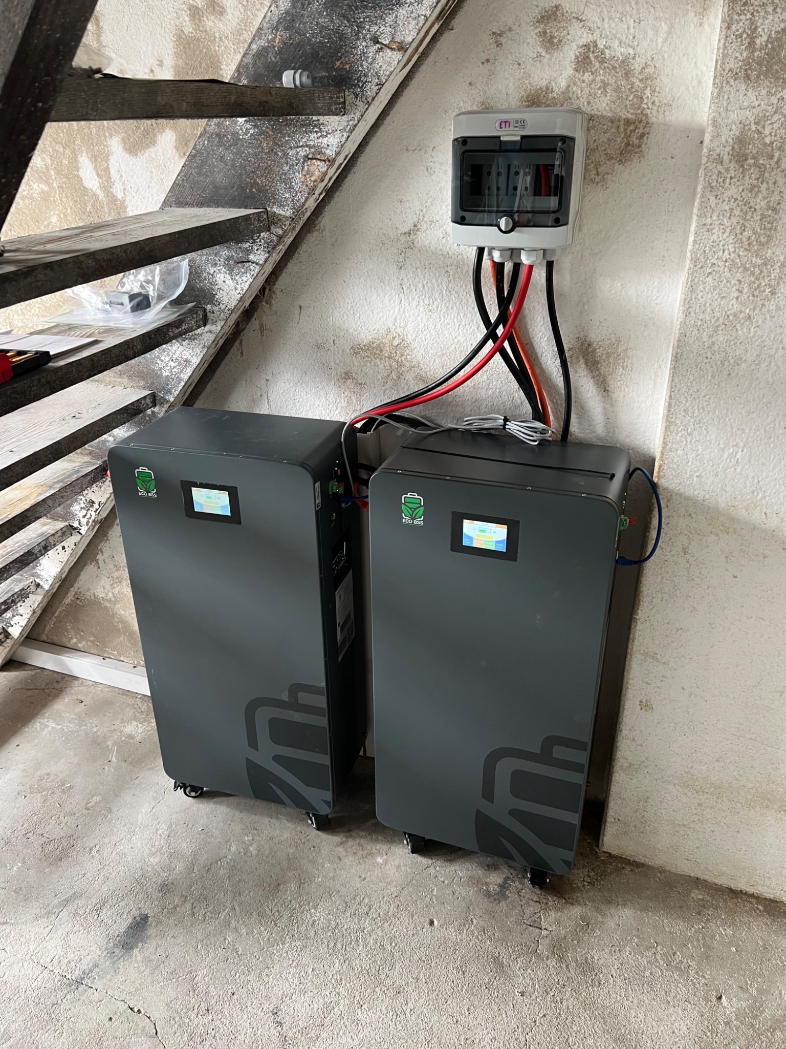 Dwa szare magazyny energii Eco BSS na kółkach, podłączone kablami do puszki elektrycznej na ścianie, umieszczone w pobliżu drewnianych schodów w surowym pomieszczeniu.
