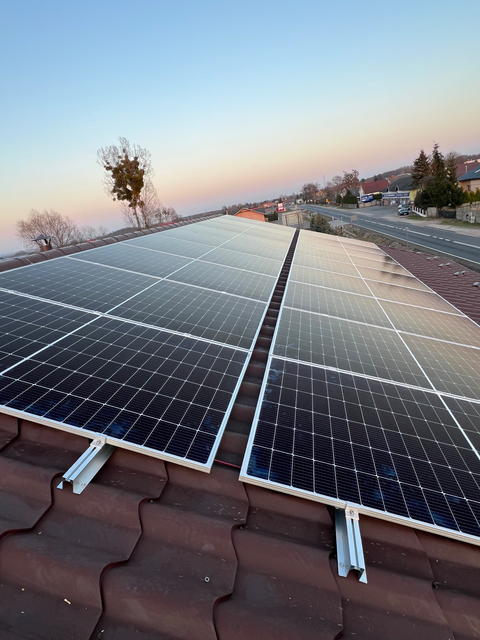Panele fotowoltaiczne na brązowym dachu, widok z góry, w tle droga i zabudowania, niebo o świcie z delikatnymi różowymi odcieniami.
