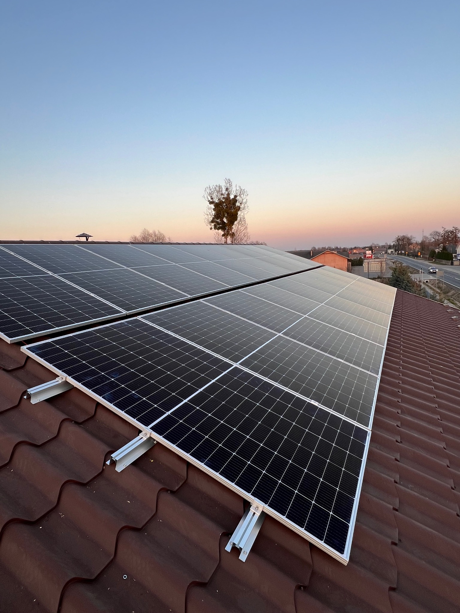 Panele fotowoltaiczne na dachu z brązowej dachówki w promieniach zachodzącego słońca. Widok na okolicę z drzewami i drogą w oddali.