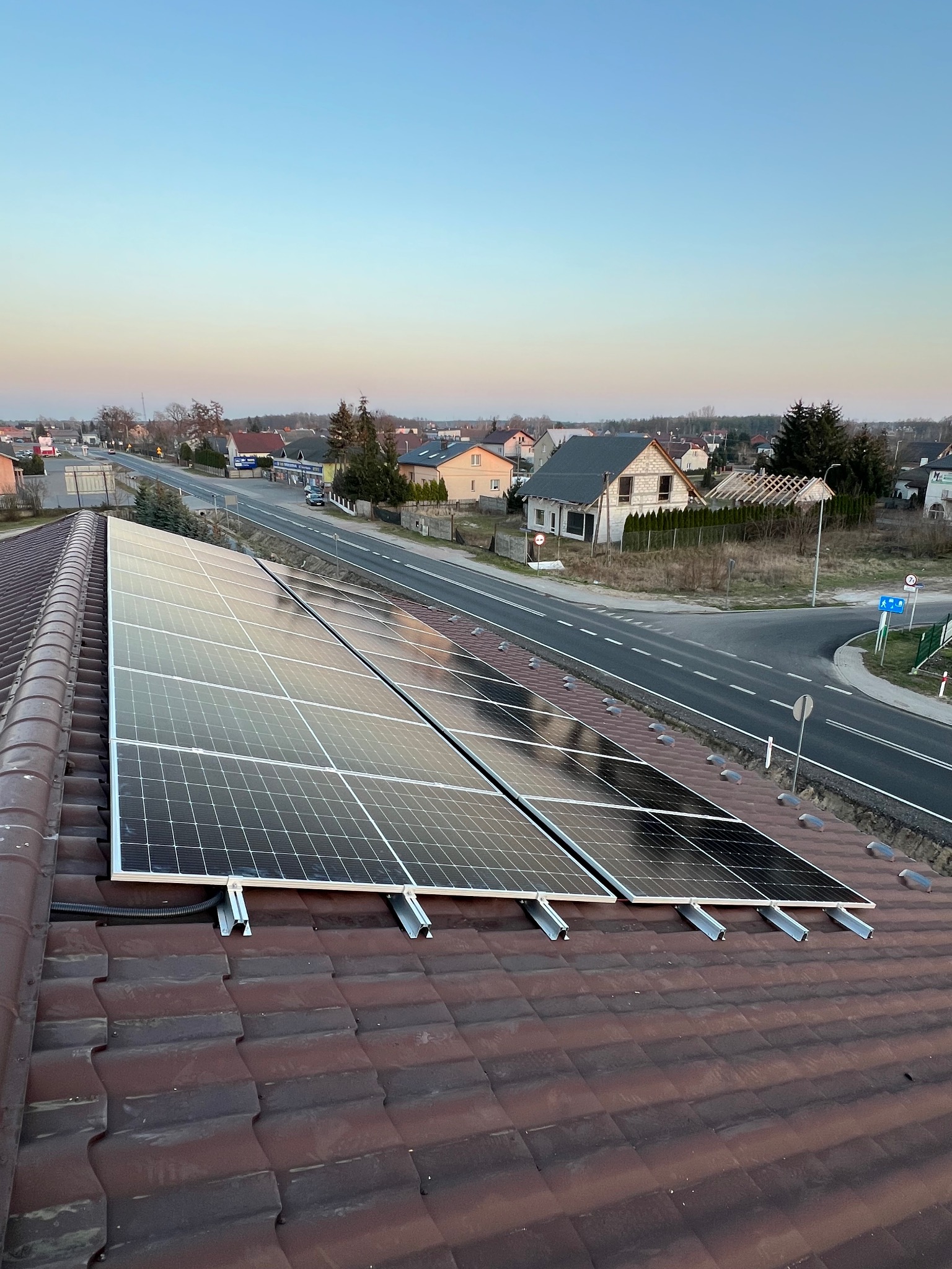 Panele fotowoltaiczne na brązowym dachu z widokiem na okolicę o zmierzchu. Widoczne metalowe mocowania i kable.