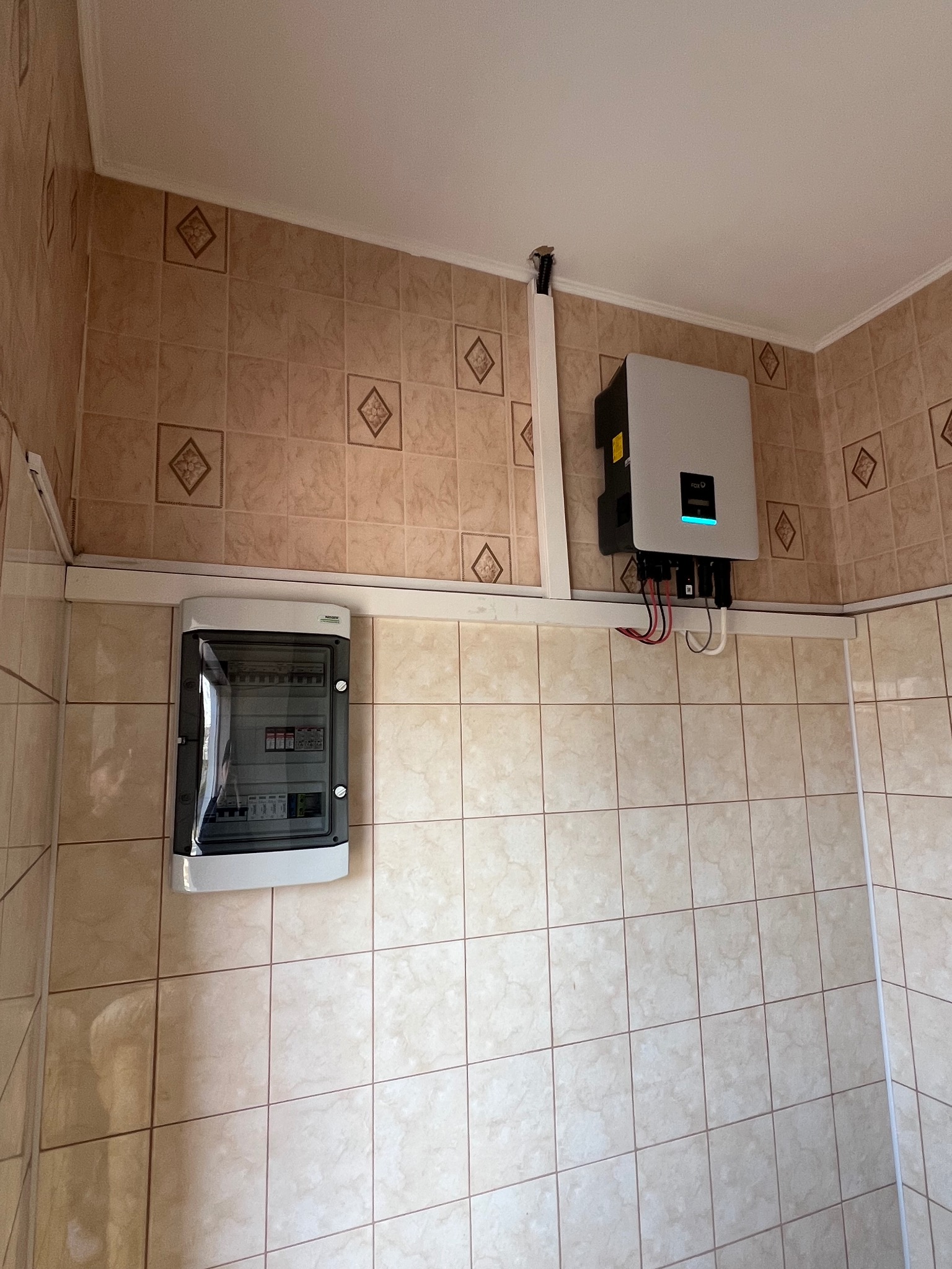 Zamontowany inwerter fotowoltaiczny z rozdzielnicą na ścianie wyłożonej beżowymi płytkami. Widoczne kable i rurki instalacyjne.
