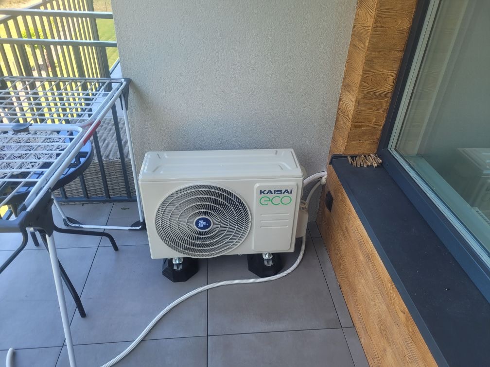 Kompaktowa jednostka zewnętrzna klimatyzacji Kaisai Eco na balkonie, umieszczona na gumowych podkładkach antywibracyjnych, z widoczną białą rurą odprowadzającą skropliny.