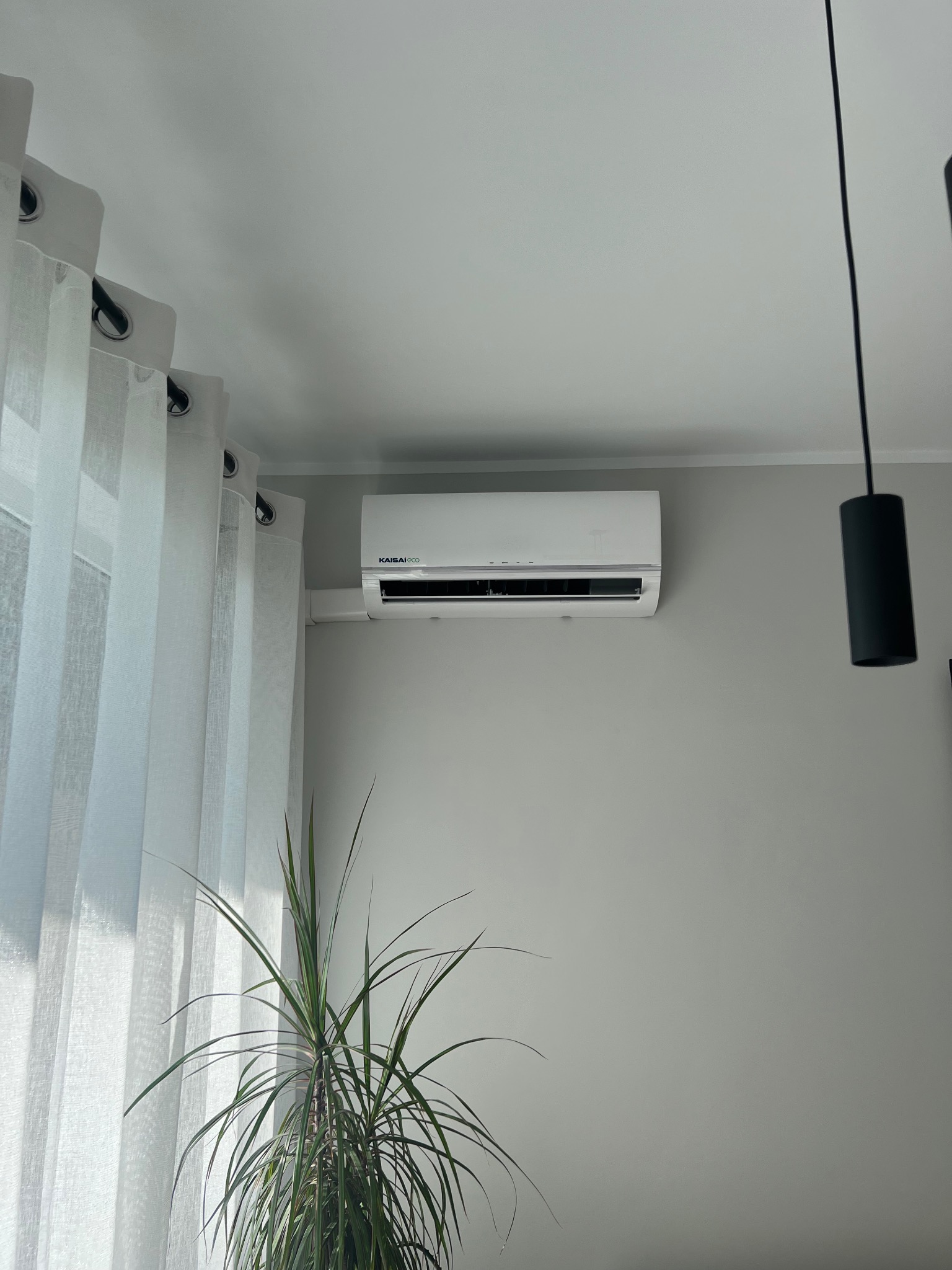 Biały klimatyzator Kaisi ECO zamontowany na szarej ścianie w jasnym wnętrzu. Obok okno z białymi zasłonami i wisząca lampa.