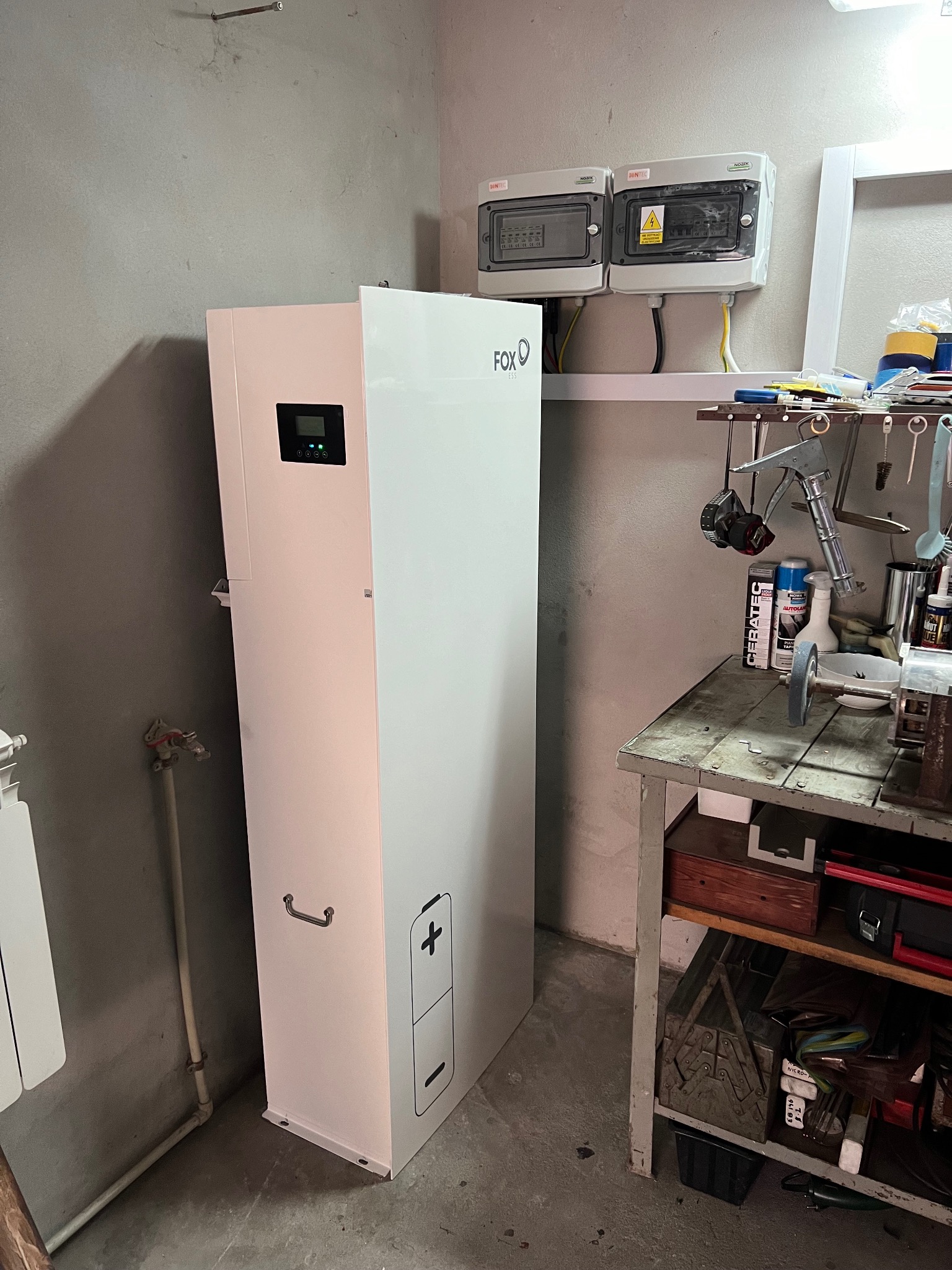 Biały magazyn energii FoxESS w pomieszczeniu z szarą ścianą, obok skrzynki bezpiecznikowe i warsztatowy stół z narzędziami. Widoczny panel sterowania i grafika baterii.