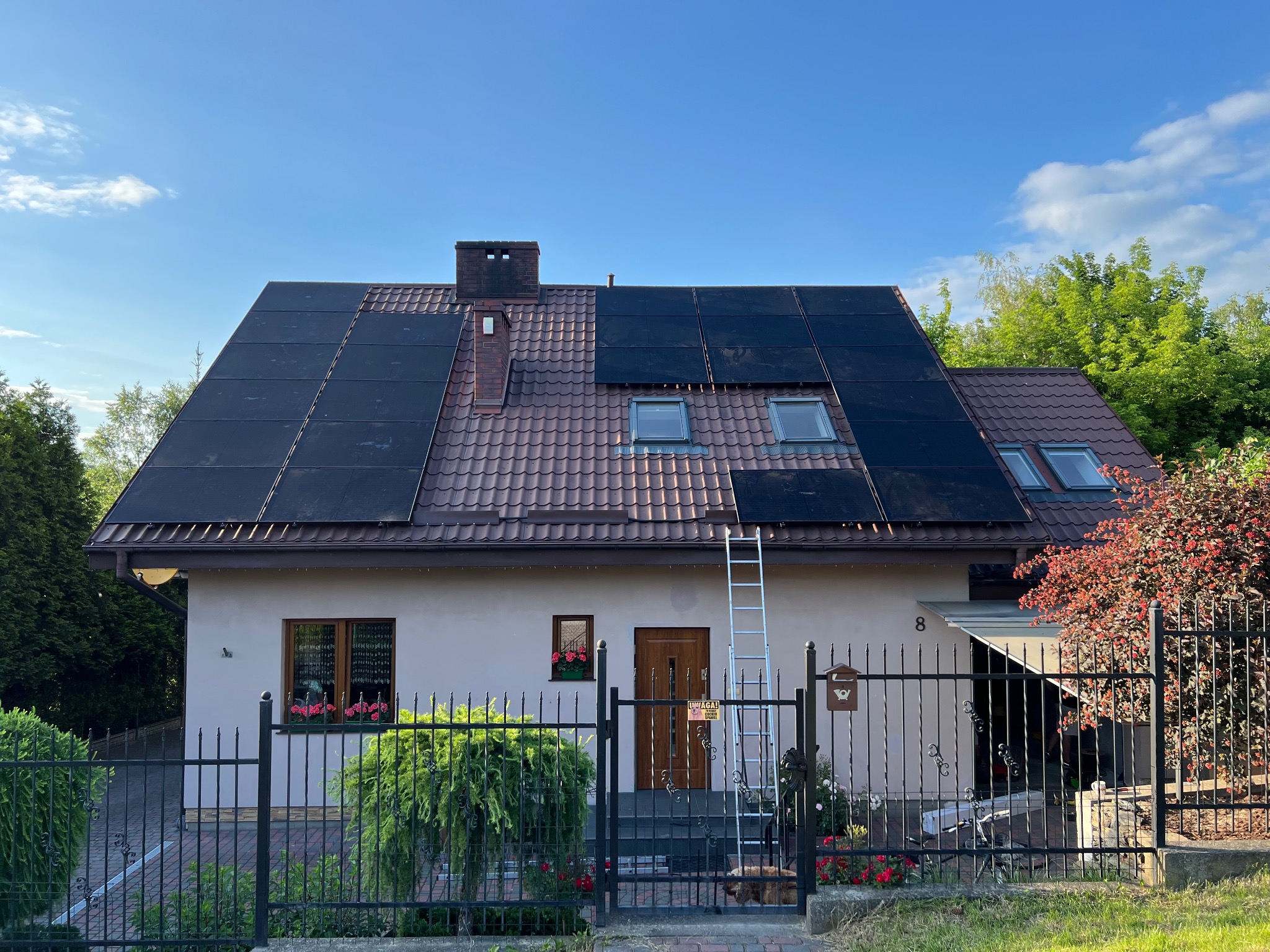 Dom jednorodzinny z częściowo zamontowanymi panelami fotowoltaicznymi na dachu z czerwoną dachówką, drabina oparta o dach, błękitne niebo.