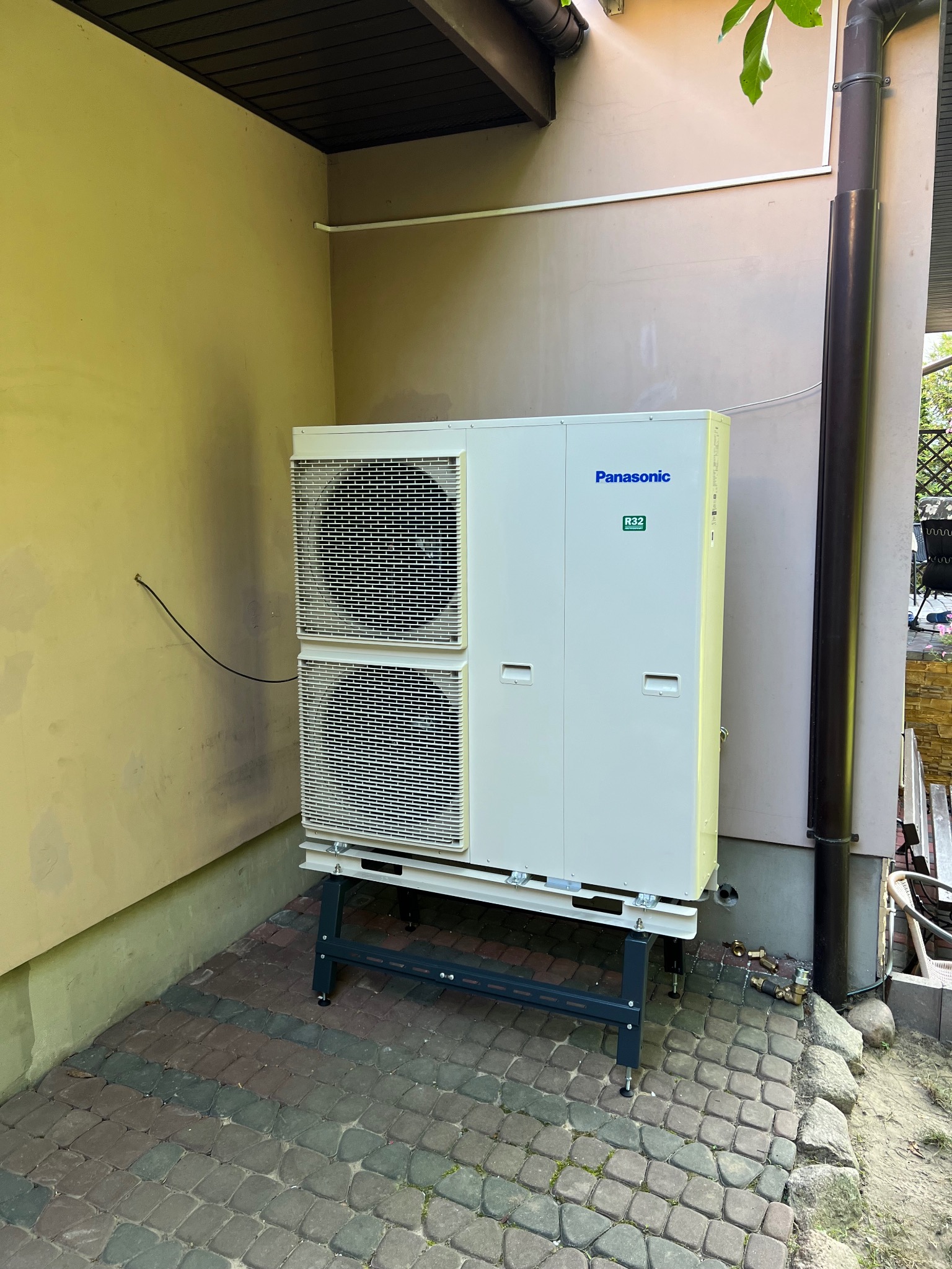 Biała pompa ciepła Panasonic R32 na zewnątrz budynku, ustawiona na czarnym stelażu, z widocznymi elementami instalacji. Obok rynna spustowa.