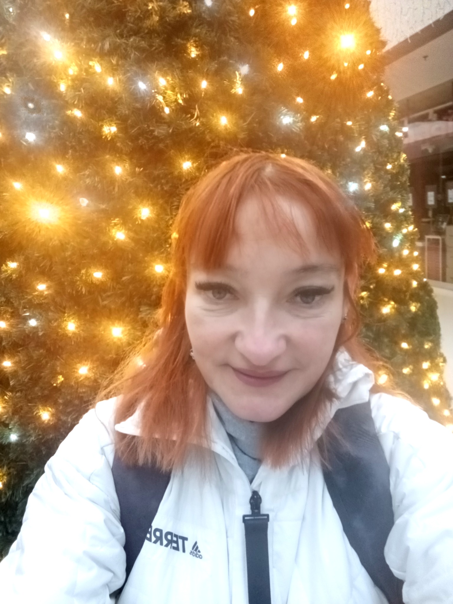 Portret kobiety z rudymi włosami na tle choinki z lampkami w centrum handlowym. Ubrana w białą kurtkę z logo. Uśmiech na twarzy, ciepłe oświetlenie.