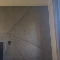 Szara ściana z geometrycznym wzorem, ciemna szafka RTV, drewniana podłoga i zasłona w rogu pokoju. Nowoczesny design wnętrza, widok na fragment salonu.