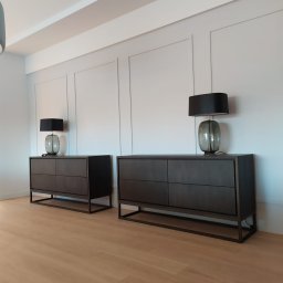 SEBASTIAN GORZOCH TopRemoncik - Minimalistyczne wnętrze: dwie ciemne komody na metalowych nogach, lampy z szarymi kloszami na tle białej ściany z dekoracyjnymi panelami i drewnianej podłogi.
