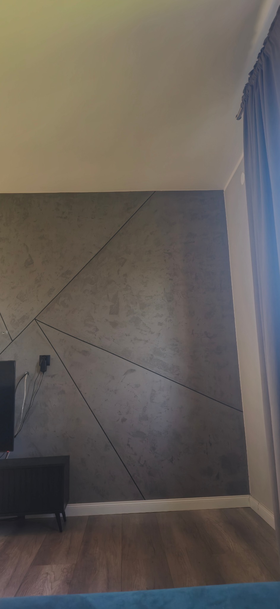 Szara ściana z geometrycznym wzorem, ciemna szafka RTV, drewniana podłoga i zasłona w rogu pokoju. Nowoczesny design wnętrza, widok na fragment salonu.