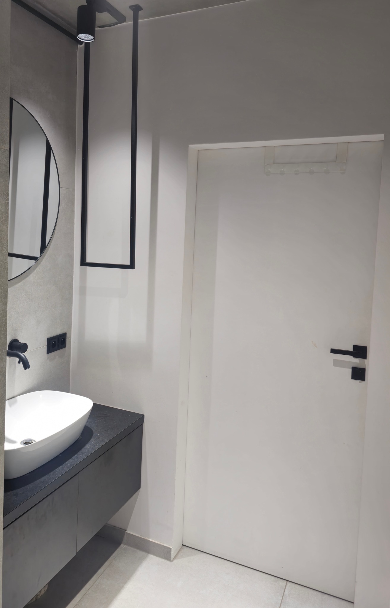 Nowoczesna łazienka z umywalką nablatową, okrągłym lustrem i czarną armaturą. Minimalistyczny design podkreślają białe ściany i drzwi oraz szara podłoga.