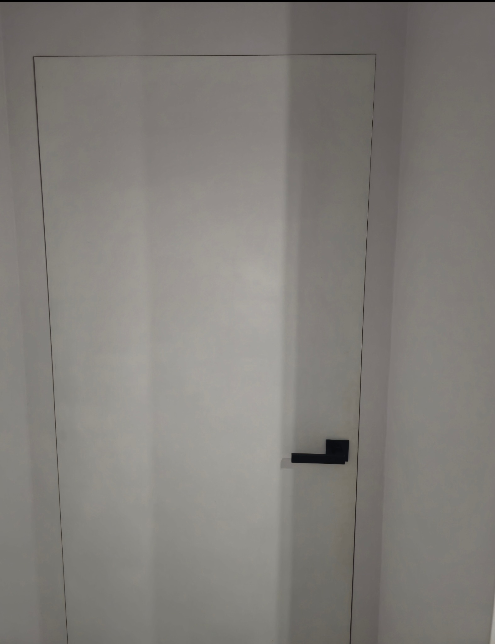 Minimalistyczne, białe drzwi wewnętrzne z czarną, prostokątną klamką. Drzwi są wbudowane w ścianę, tworząc jednolitą powierzchnię. Gładka faktura i proste linie.