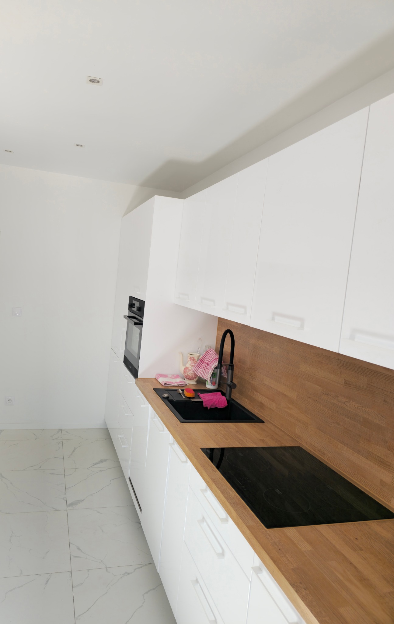 Nowoczesna kuchnia z białymi szafkami, drewnianym blatem i czarnym zlewem oraz płytą indukcyjną. Marmurowa podłoga, minimalistyczny design.