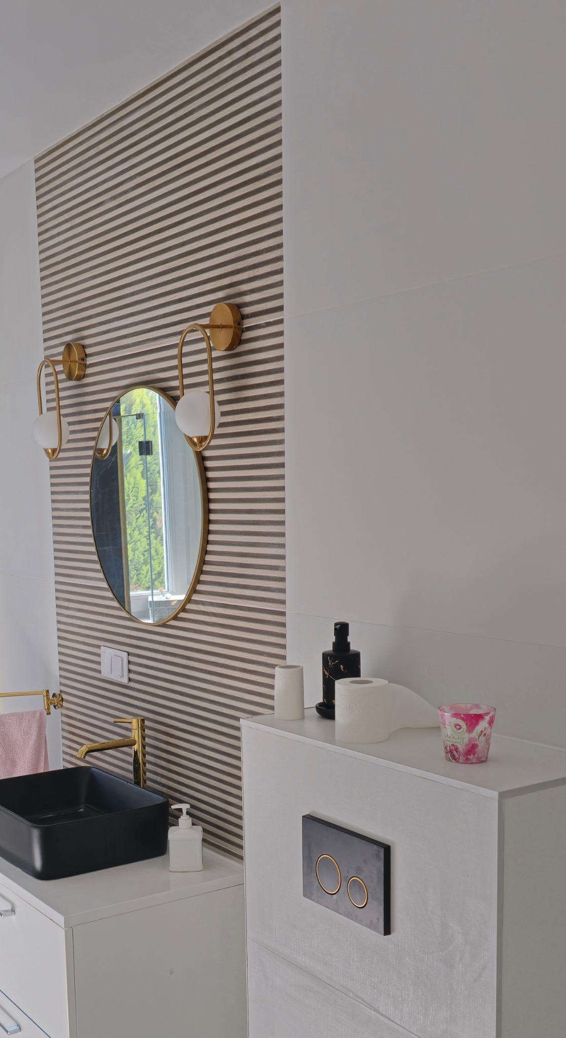 Nowoczesna łazienka z czarną umywalką nablatową, złotą armaturą i okrągłym lustrem na tle ściany w poziome pasy. Minimalistyczny design, eleganckie oświetlenie.