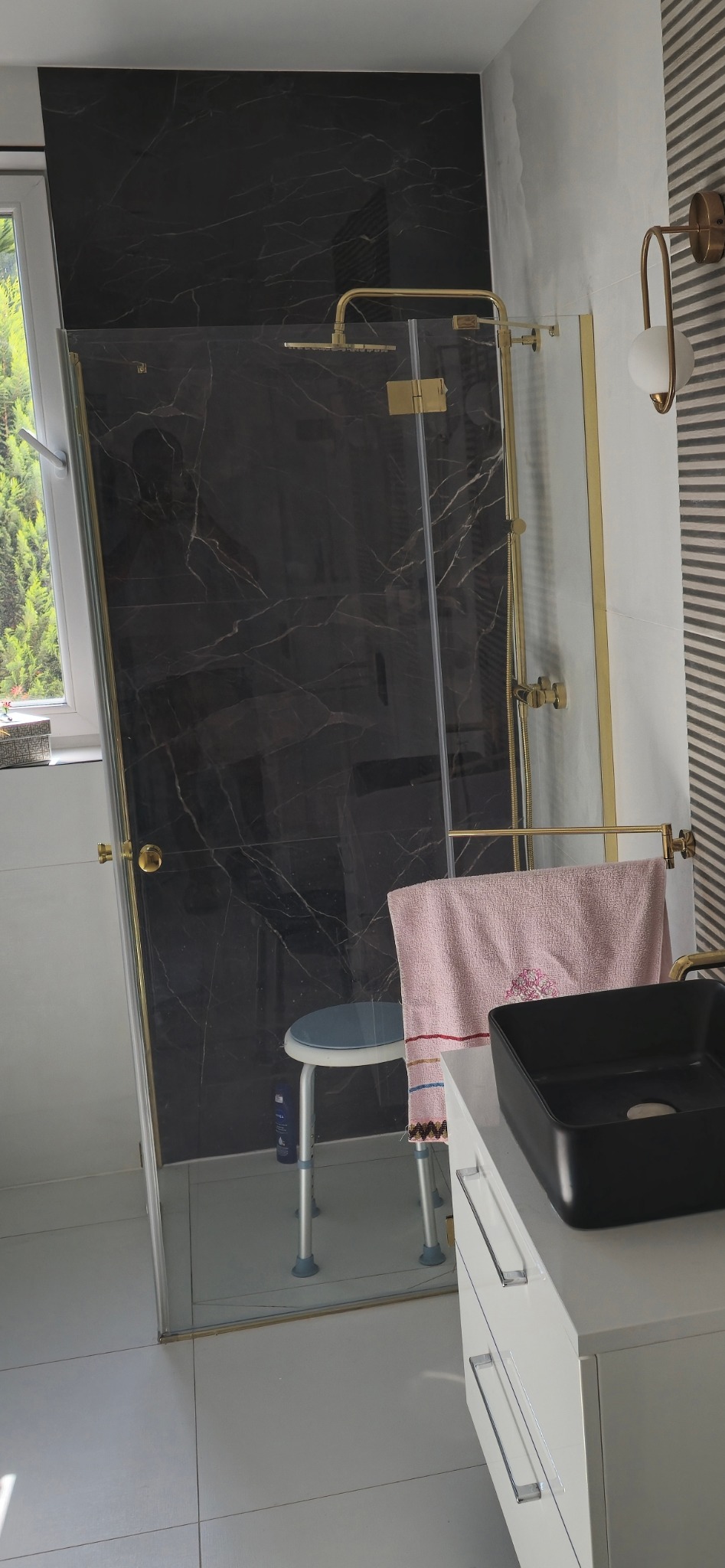 Nowoczesna kabina prysznicowa z czarnym marmurem i złotymi detalami, widoczna z umywalką i szafką w łazience po remoncie. Krzesło prysznicowe w tle.