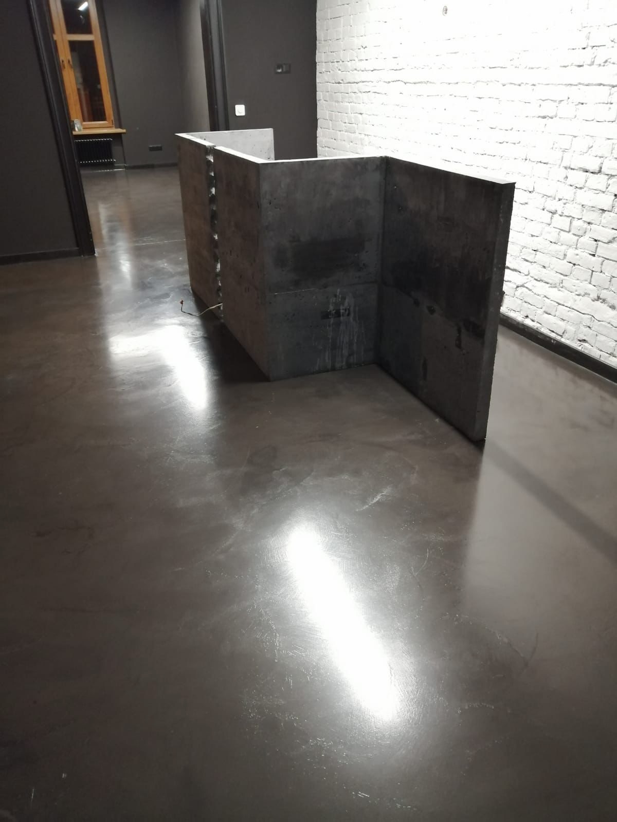 Betonowa konstrukcja w surowym wnętrzu. Szara podłoga z połyskiem odbija światło. Biała ściana z cegły w tle. Minimalistyczny design.