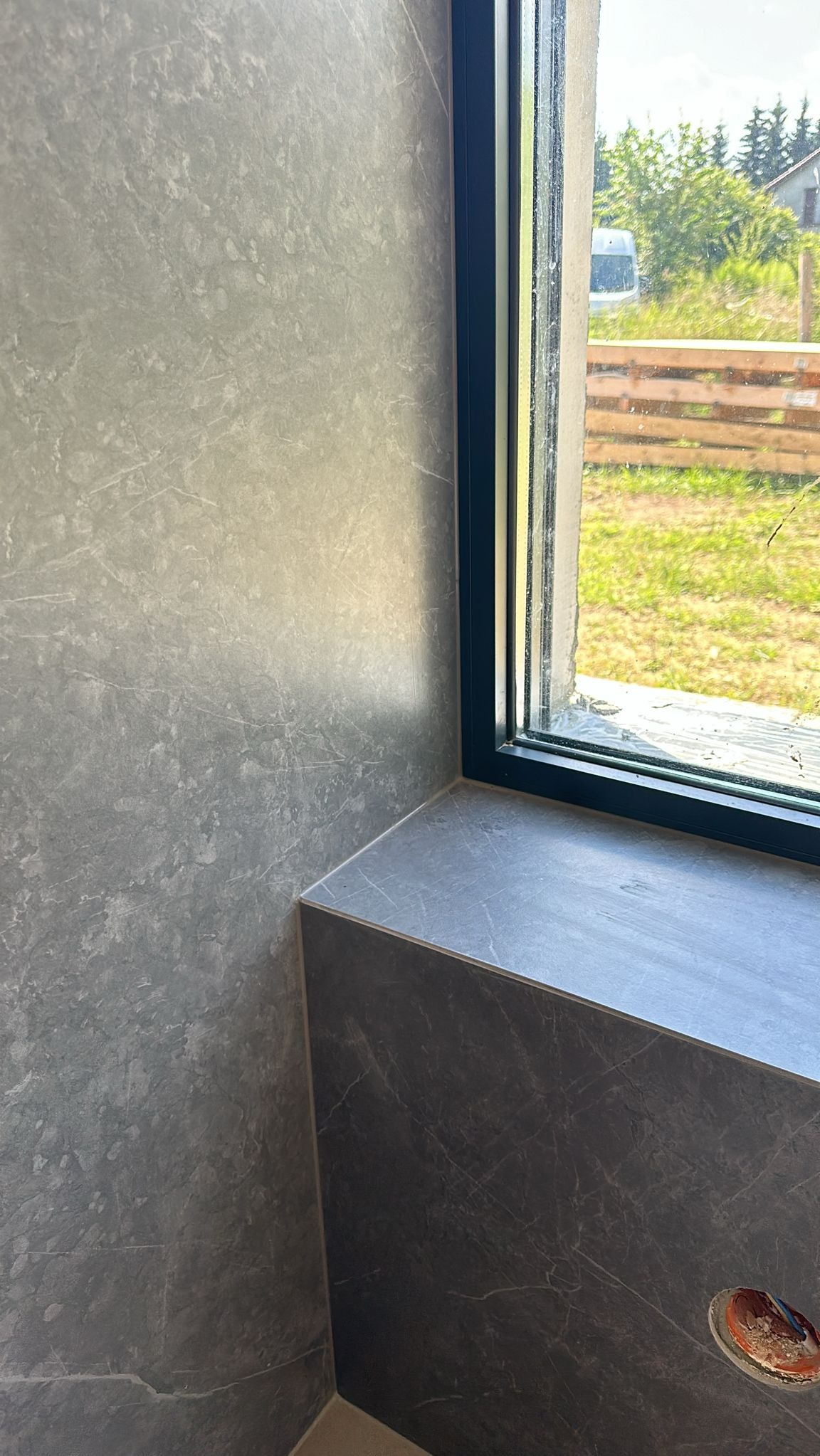 Szara, marmurkowa okładzina ściany i parapetu przy oknie z czarną ramą. Widoczny fragment instalacji elektrycznej. Na zewnątrz zieleń i drewniany płot.