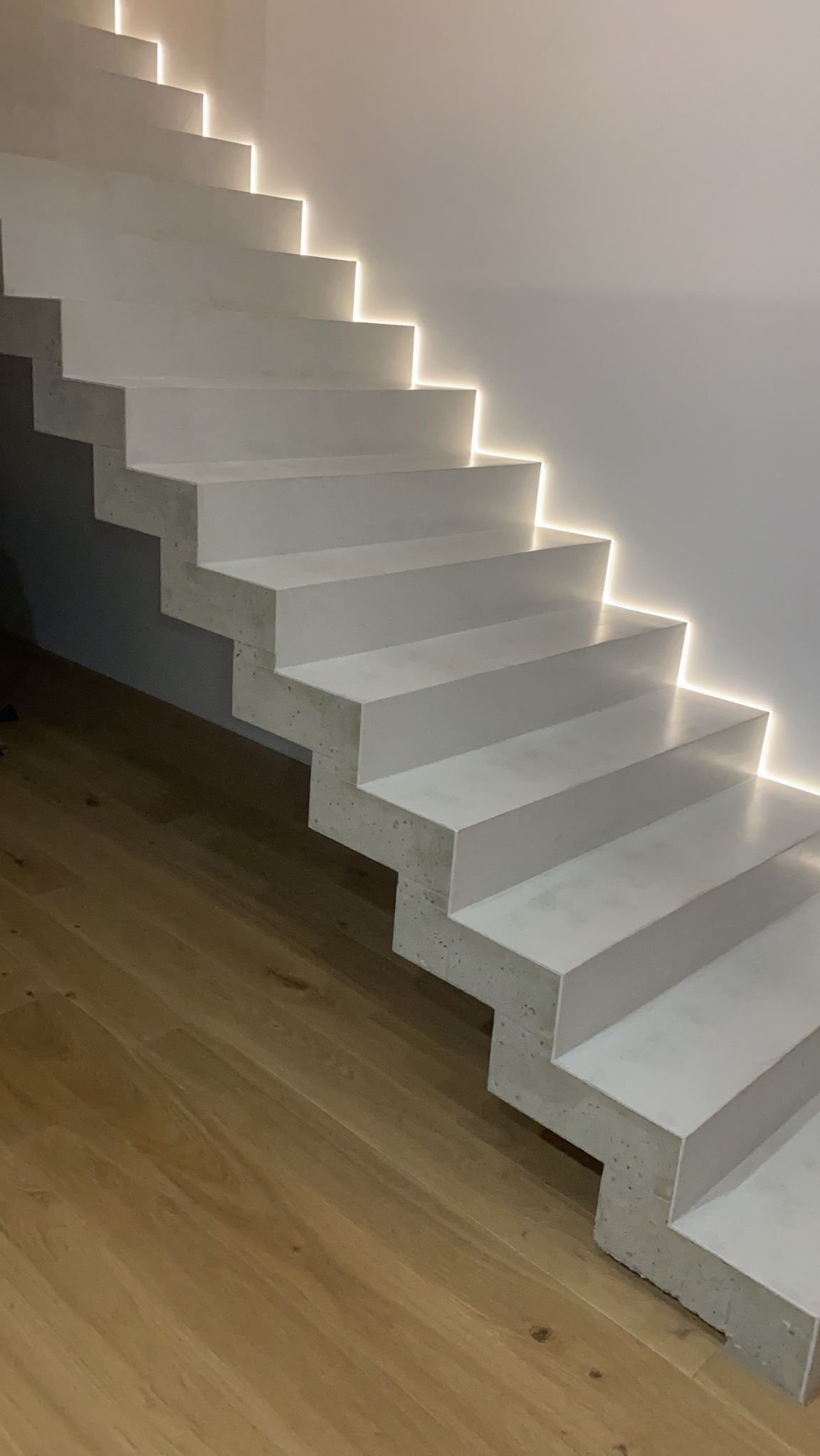 Betonowe schody z subtelnym oświetleniem LED, minimalistyczny design. Jasne drewno podłogi kontrastuje z surowością betonu. Nowoczesne wykończenie wnętrza.
