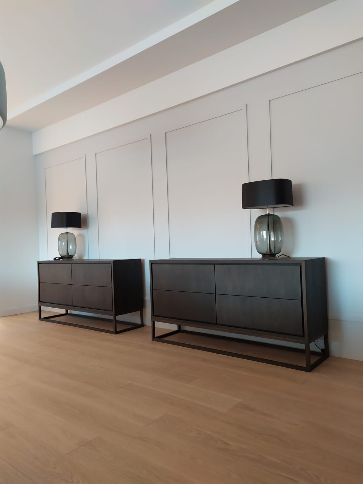 Minimalistyczne wnętrze: dwie ciemne komody na metalowych nogach, lampy z szarymi kloszami na tle białej ściany z dekoracyjnymi panelami i drewnianej podłogi.
