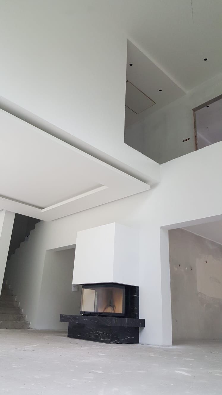 Nowoczesny salon w trakcie wykańczania: białe ściany, sufit z podwieszanymi elementami, czarny kominek i schody. Widoczne ślady prac wykończeniowych.