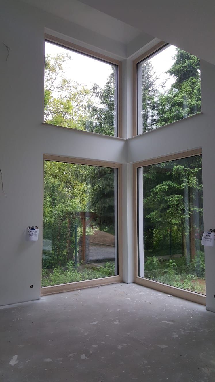 Wnętrze pokoju z trzema dużymi oknami z drewnianymi ramami, przez które widać zieleń drzew. Ściany białe, podłoga szara, surowa. Widoczne gniazdka elektryczne.