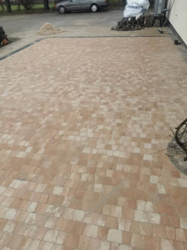 Ułożona kostka brukowa w odcieniach beżu i brązu tworzy wzór na podjeździe. Widoczne są materiały budowlane i fragment samochodu w tle.