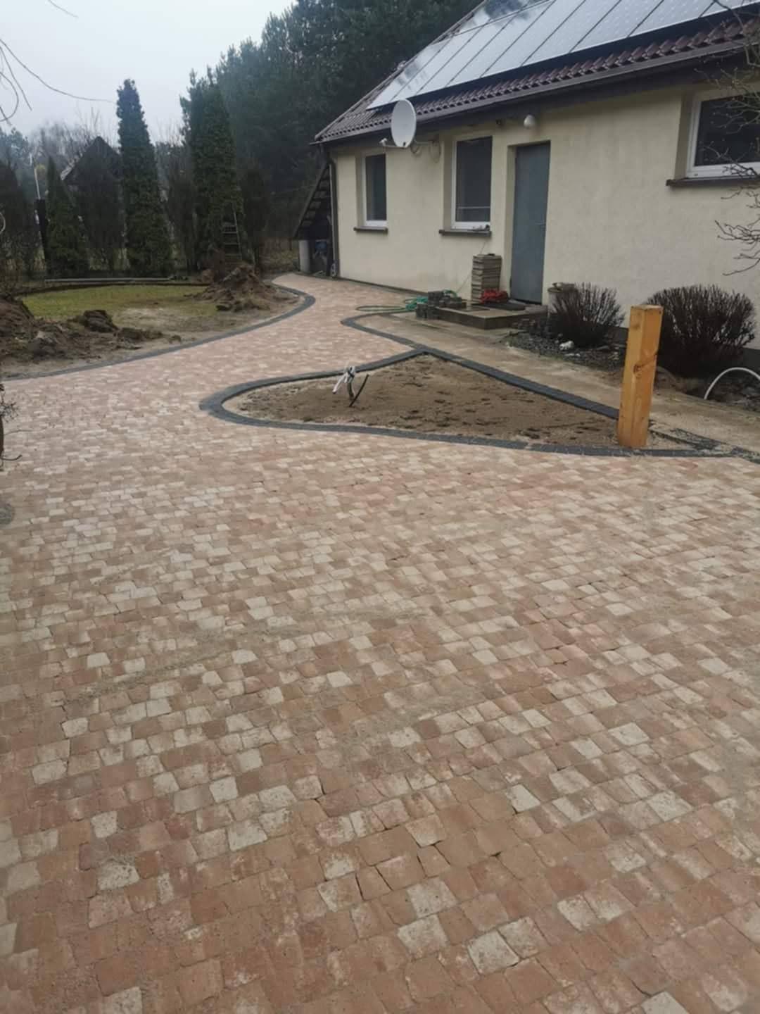 Kaskadowa ścieżka z czerwonej kostki brukowej, obramowana ciemnym pasem, prowadzi do domu z panelami słonecznymi. Część terenu przygotowana pod nasadzenia.