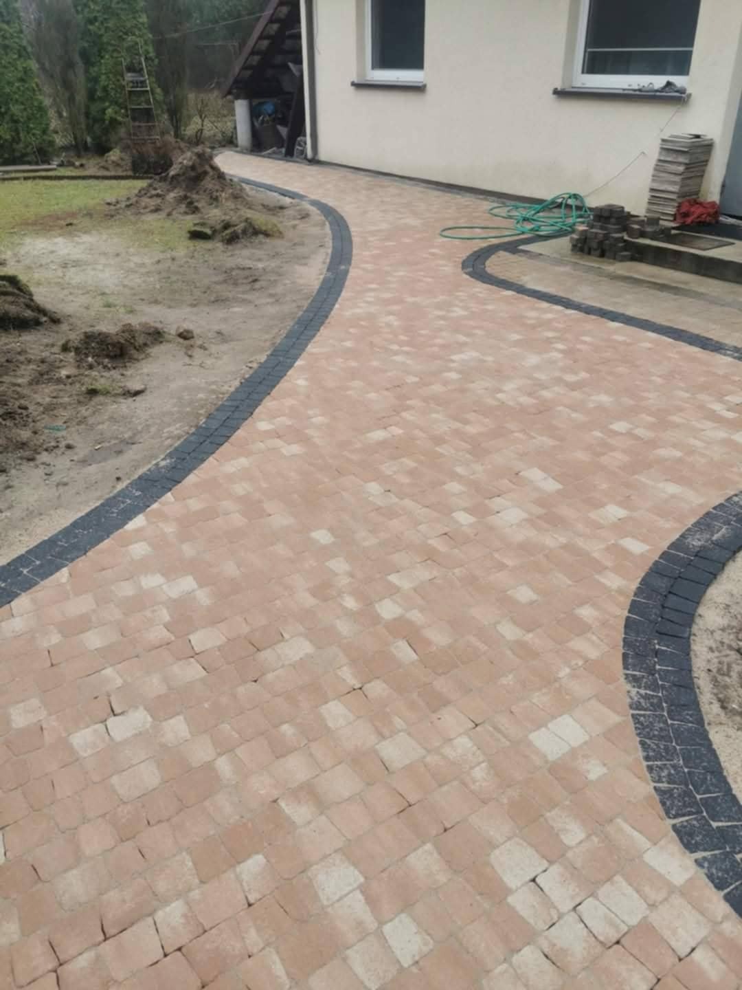 Kaskadowa ścieżka z beżowej kostki brukowej, obramowana ciemną kostką, prowadzi wzdłuż jasnego budynku. Widoczne pozostałości ziemi i narzędzia.