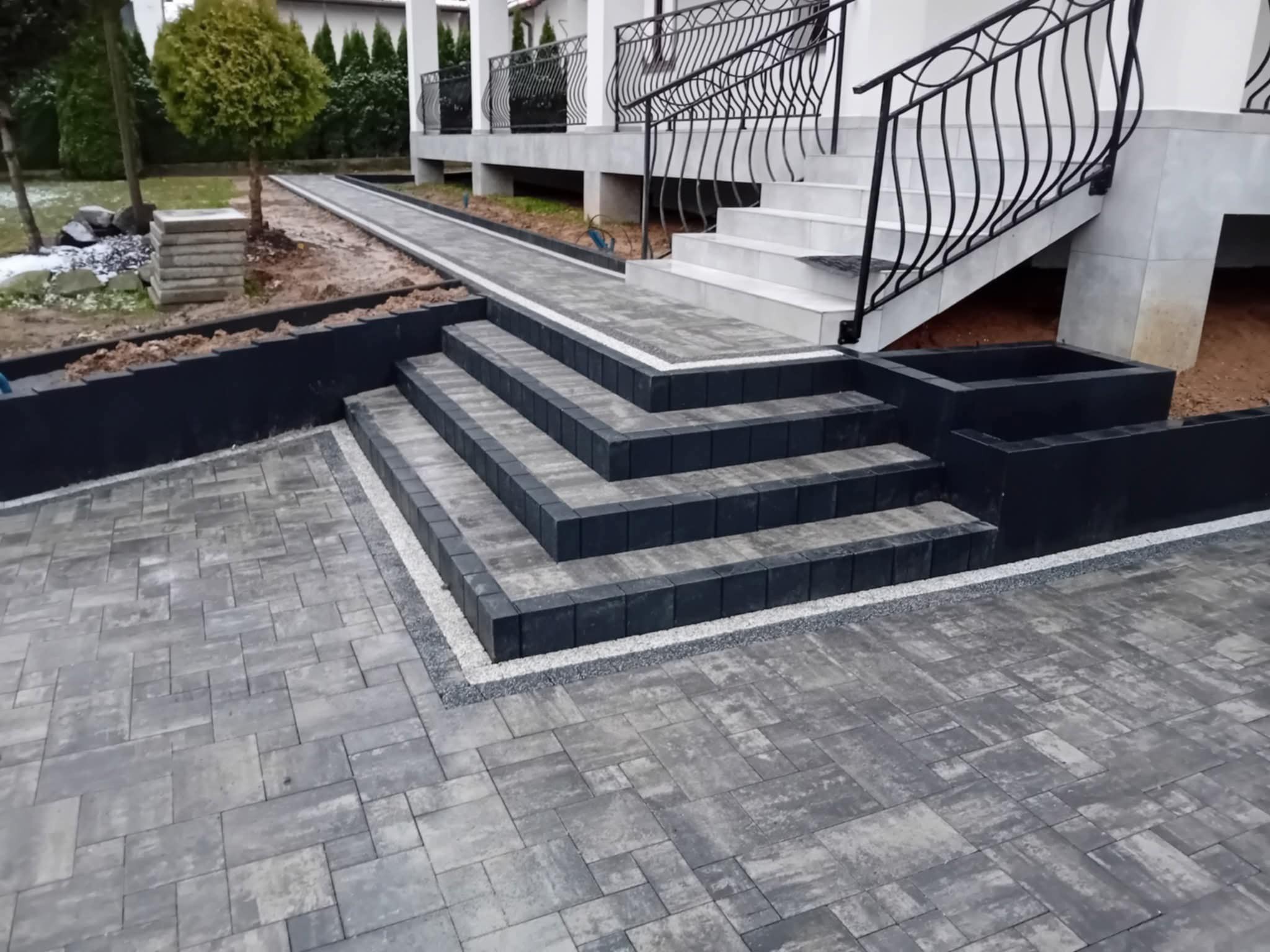 Nowoczesne schody z kostki brukowej w odcieniach szarości, z subtelnym obramowaniem z drobnych kamyczków, prowadzące do wejścia domu z balustradą.
