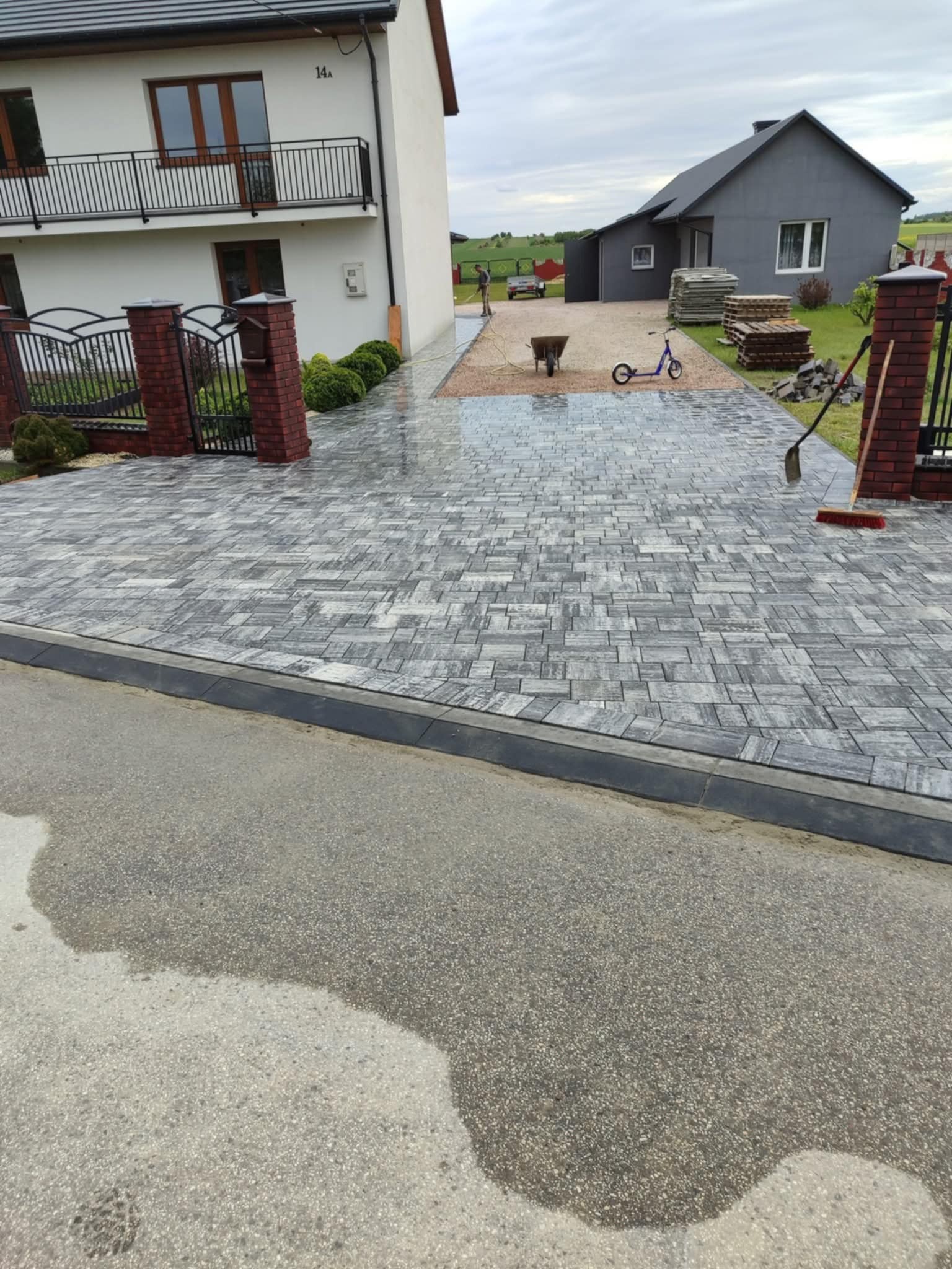 Ułożona szara kostka brukowa tworząca podjazd do domu z bramą i ogrodzeniem. W tle widać drugi budynek i przygotowane materiały budowlane. Na podjeździe stoi hulajnoga.