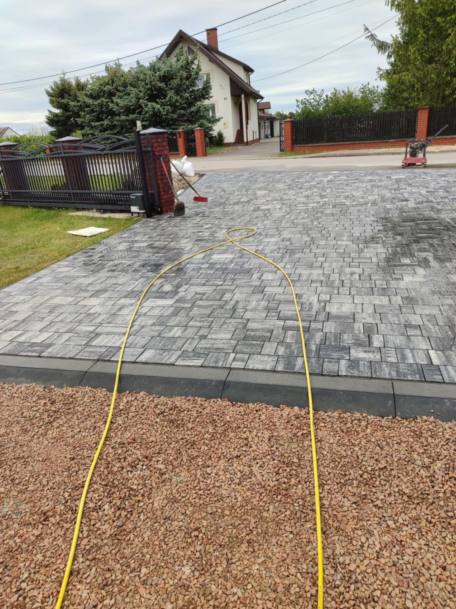 Świeżo ułożona kostka brukowa w szaro-białej tonacji na podjeździe, obok widoczny brązowy żwir i żółty wąż ogrodowy. W tle dom z ceglanym kominem i czarne ogrodzenie.