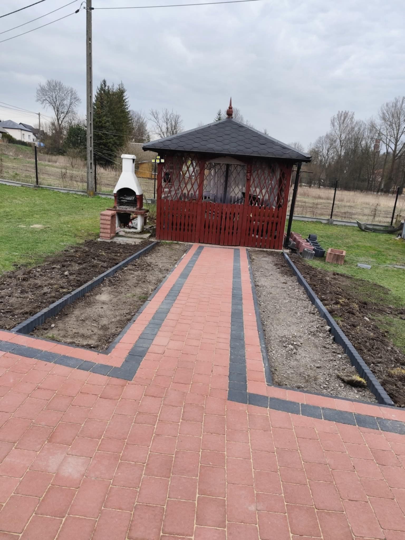 Czerwono-szara kostka brukowa prowadzi do altany i grilla. Obrzeża rabat z kostki. Częściowo zagospodarowany teren zielony. Pochmurne niebo w tle.