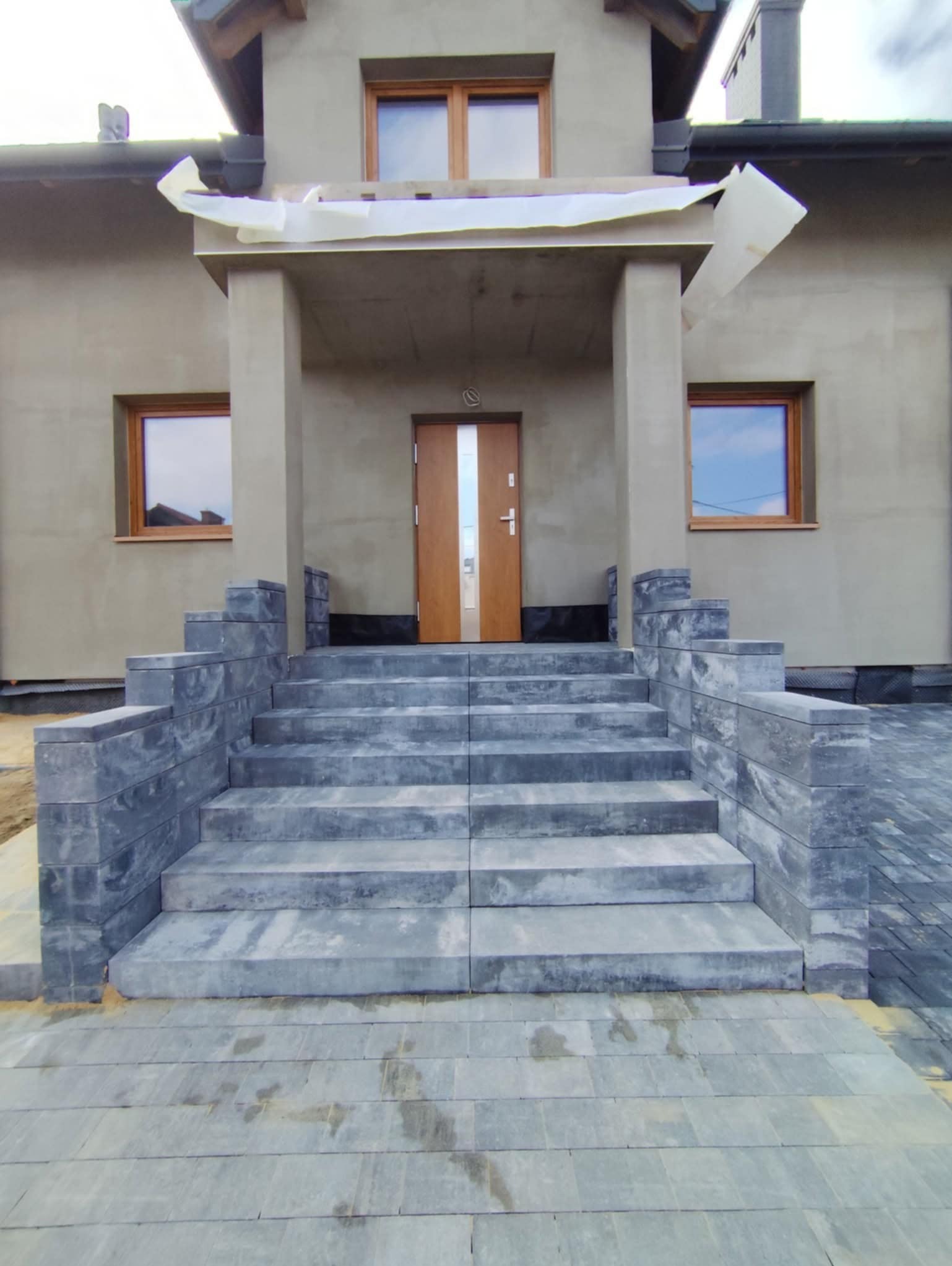 Nowe schody z kostki brukowej prowadzące do domu z drewnianymi oknami. Widoczny fragment chodnika z kostki brukowej, podkreślający estetykę wykończenia.