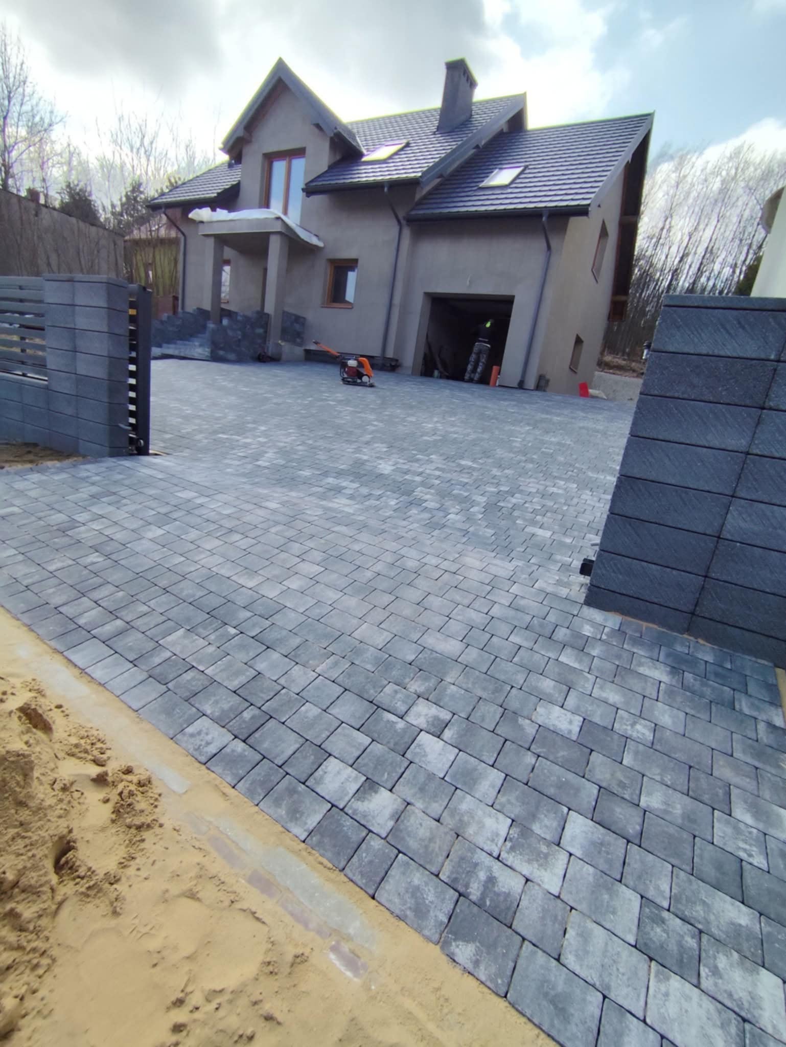 Nowoczesny podjazd z szarej kostki brukowej przed domem z garażem. Widoczny fragment ogrodzenia z betonowych bloczków i zagęszczarka w tle.