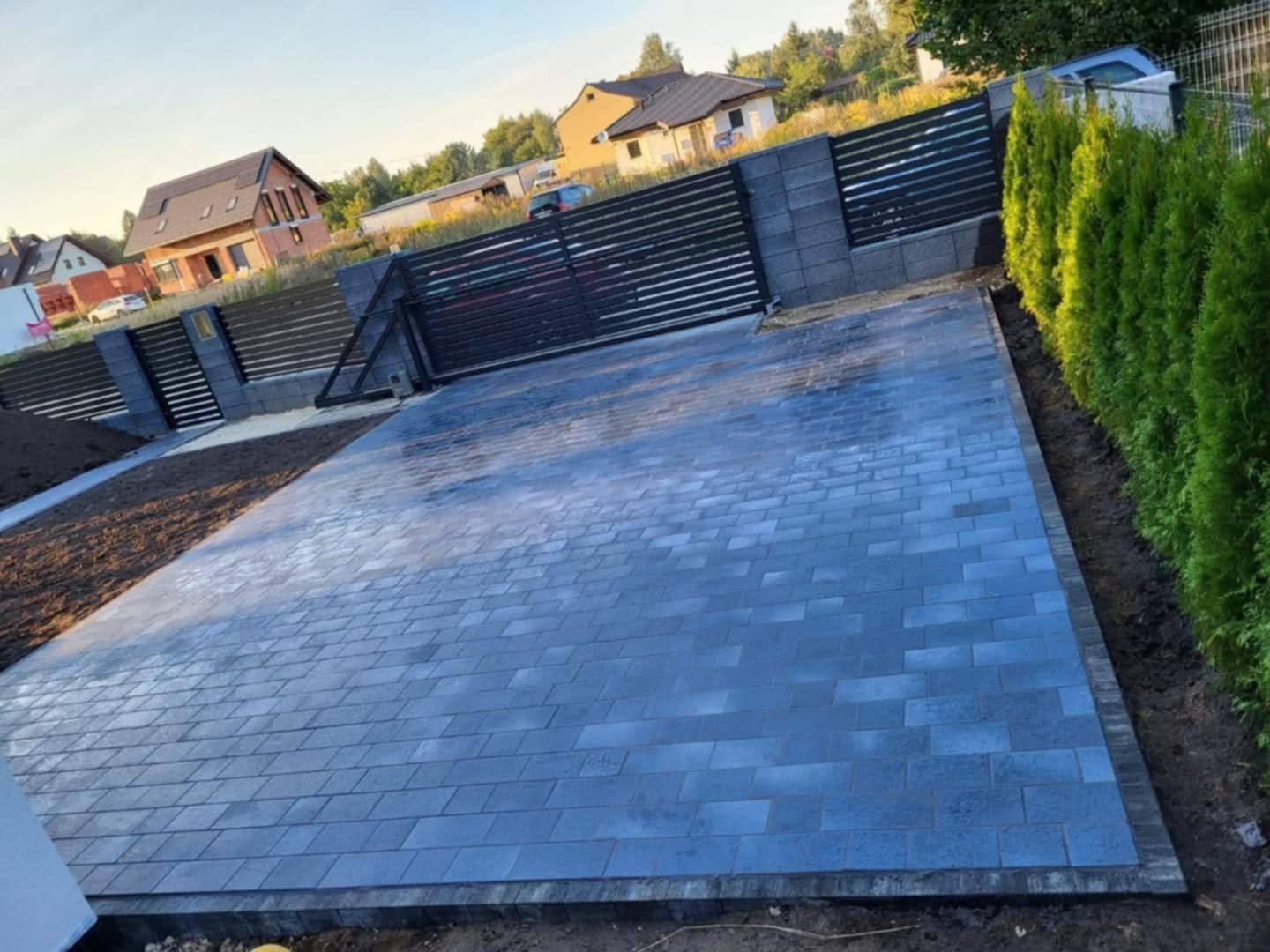 Ułożona, szara kostka brukowa na podjeździe w Zgierzu. Widoczny fragment ogrodzenia z metalowymi przęsłami i rośliny ozdobne wzdłuż granicy działki. Ukończona realizacja.
