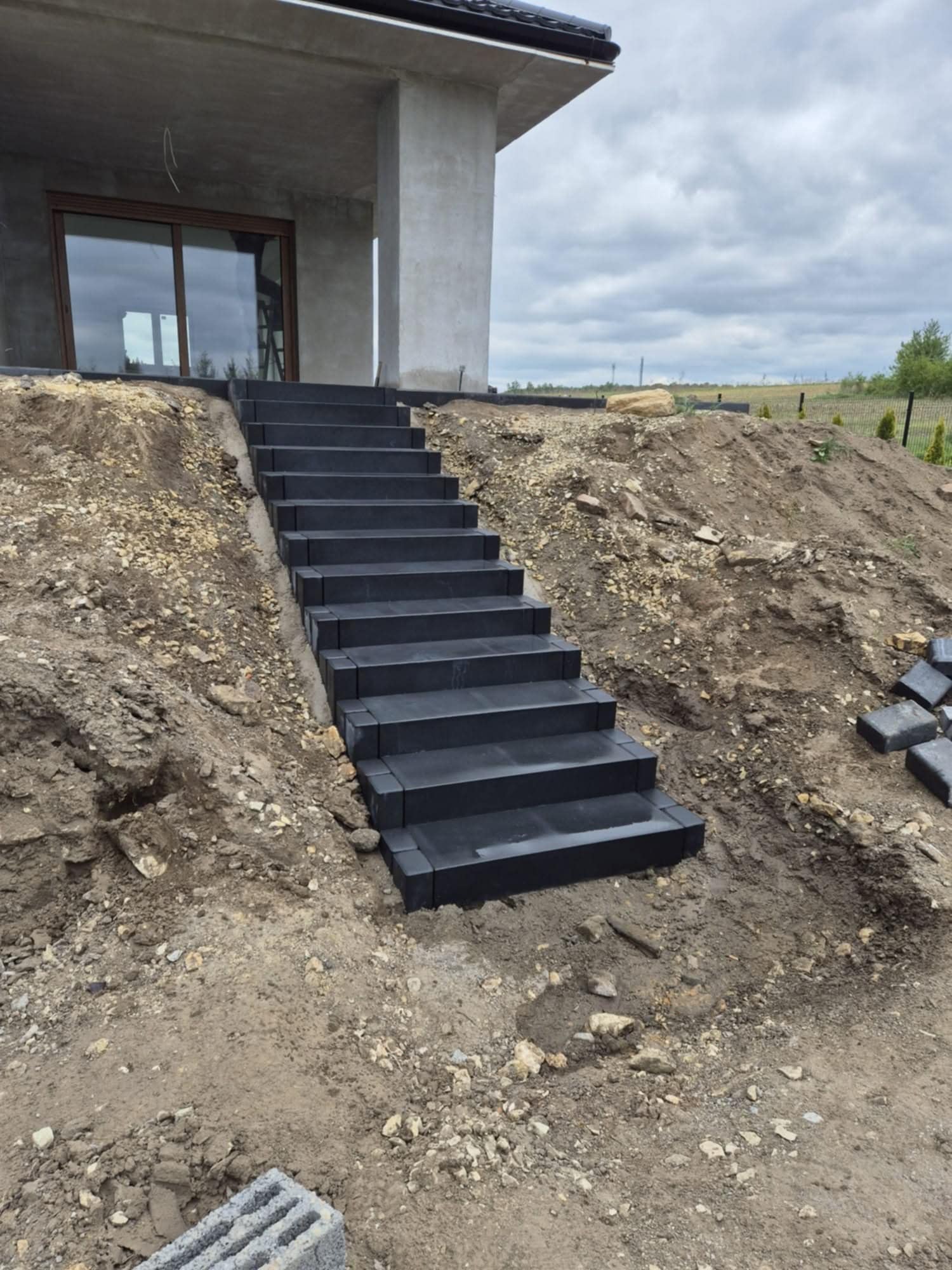 Nowoczesne, czarne schody z kostki brukowej prowadzące do domu na nierównym terenie. Widoczne niedokończone prace ziemne i kostka brukowa obok schodów.