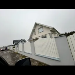 Nowoczesne ogrodzenie panelowe z białymi przęsłami i szarymi słupkami, widok z perspektywy pojazdu, dom w tle, pochmurny dzień.