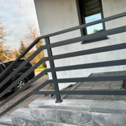 Nowoczesna, czarna balustrada metalowa z poziomymi szczeblami, zamontowana przy schodach wejściowych do domu. Widoczny fragment samochodu i otoczenie domu.