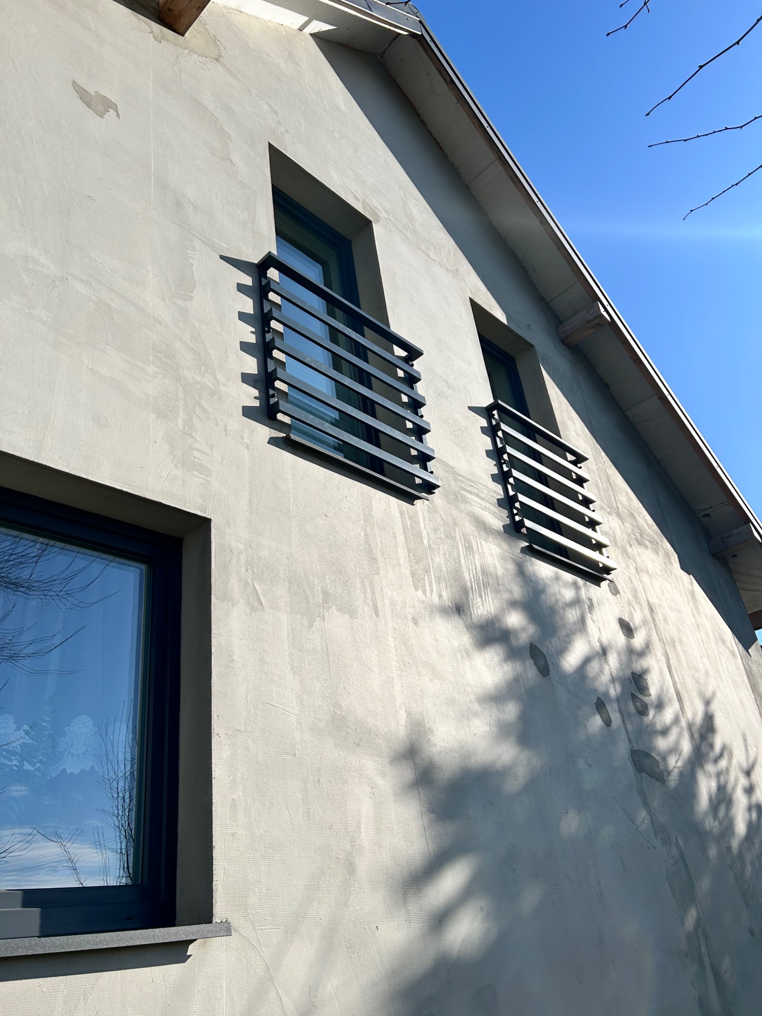 Elewacja domu z zamontowanymi, nowoczesnymi, metalowymi balustradami okiennymi w kolorze grafitowym na tle błękitnego nieba i surowej elewacji budynku.