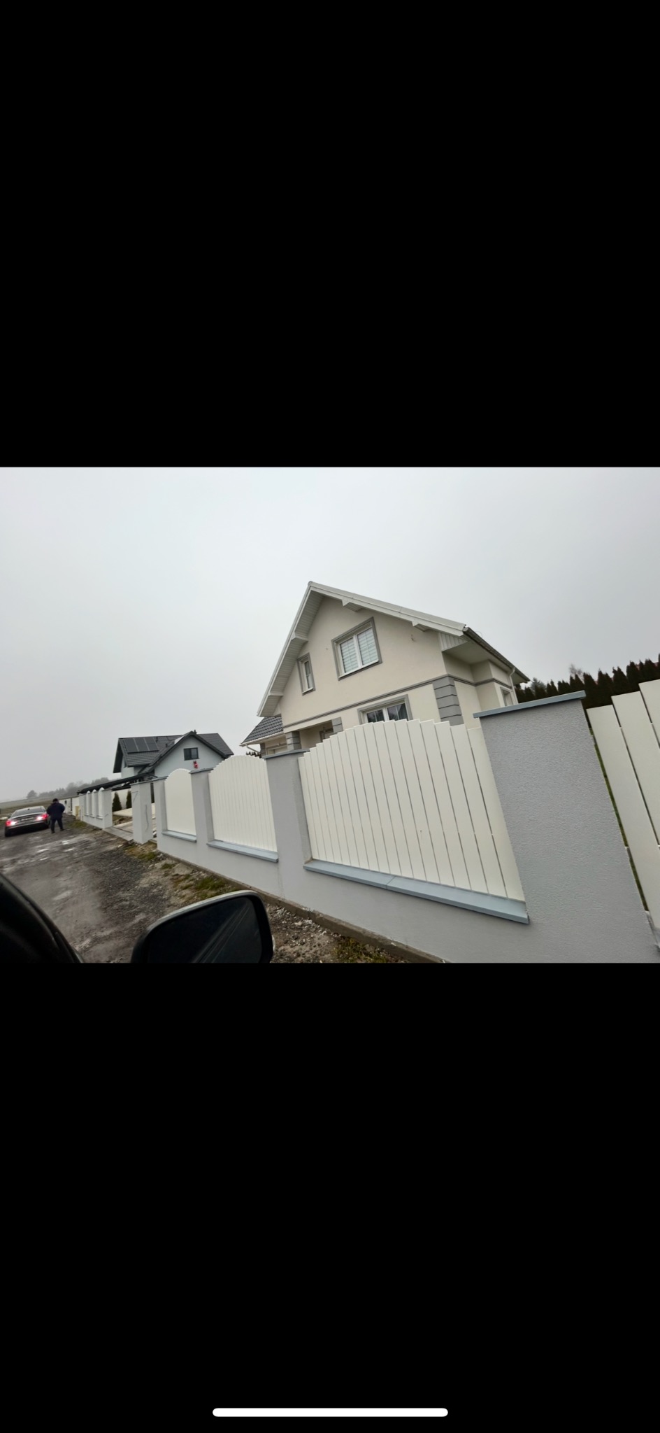 Nowoczesne ogrodzenie panelowe z białymi przęsłami i szarymi słupkami, widok z perspektywy pojazdu, dom w tle, pochmurny dzień.